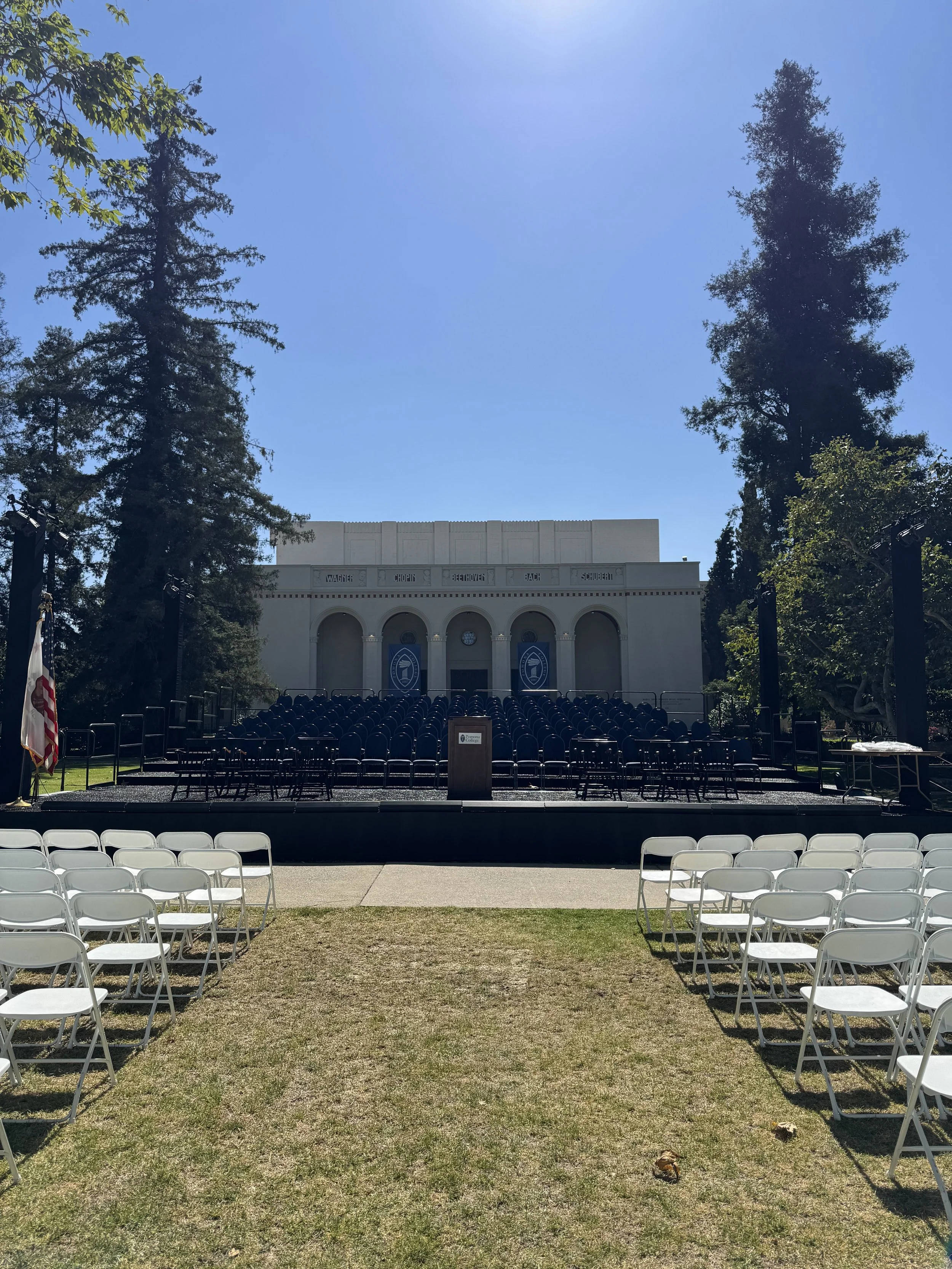 Outdoor stage setup with rows of white chairs facing a platform in front of a white building, surrounded by trees under a clear blue sky.
