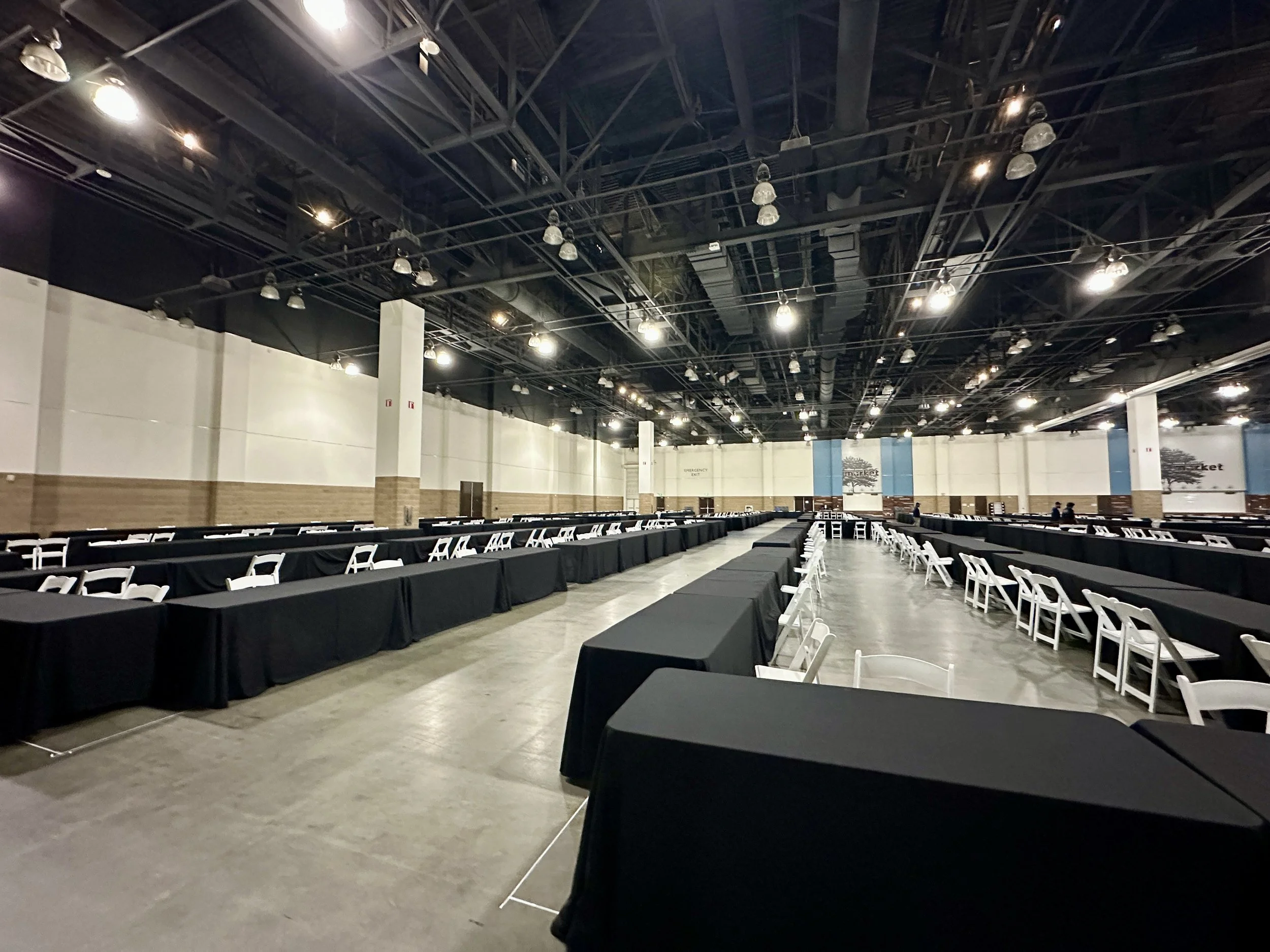Large, empty conference room with long black tables and white chairs arranged along the sides, overhead industrial lighting, and high exposed ceilings.