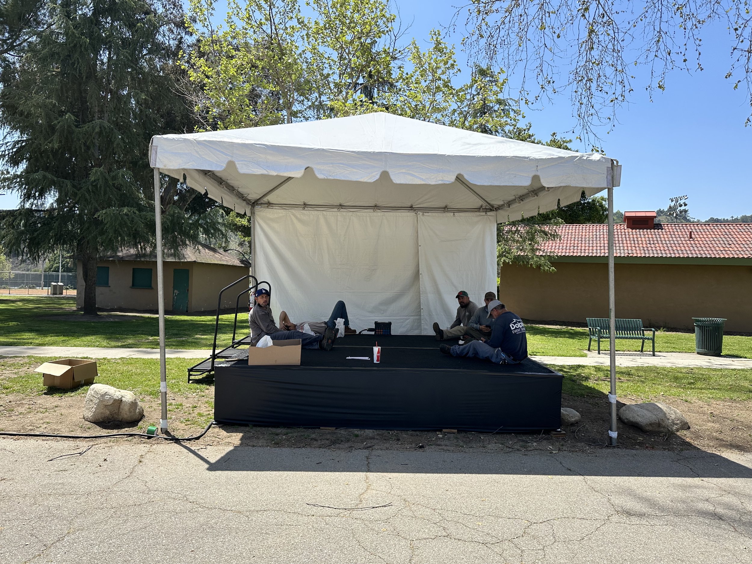 A group of people sitting and resting on a small outdoor stage under a white canopy tent in a park. There are trees and a building in the background.