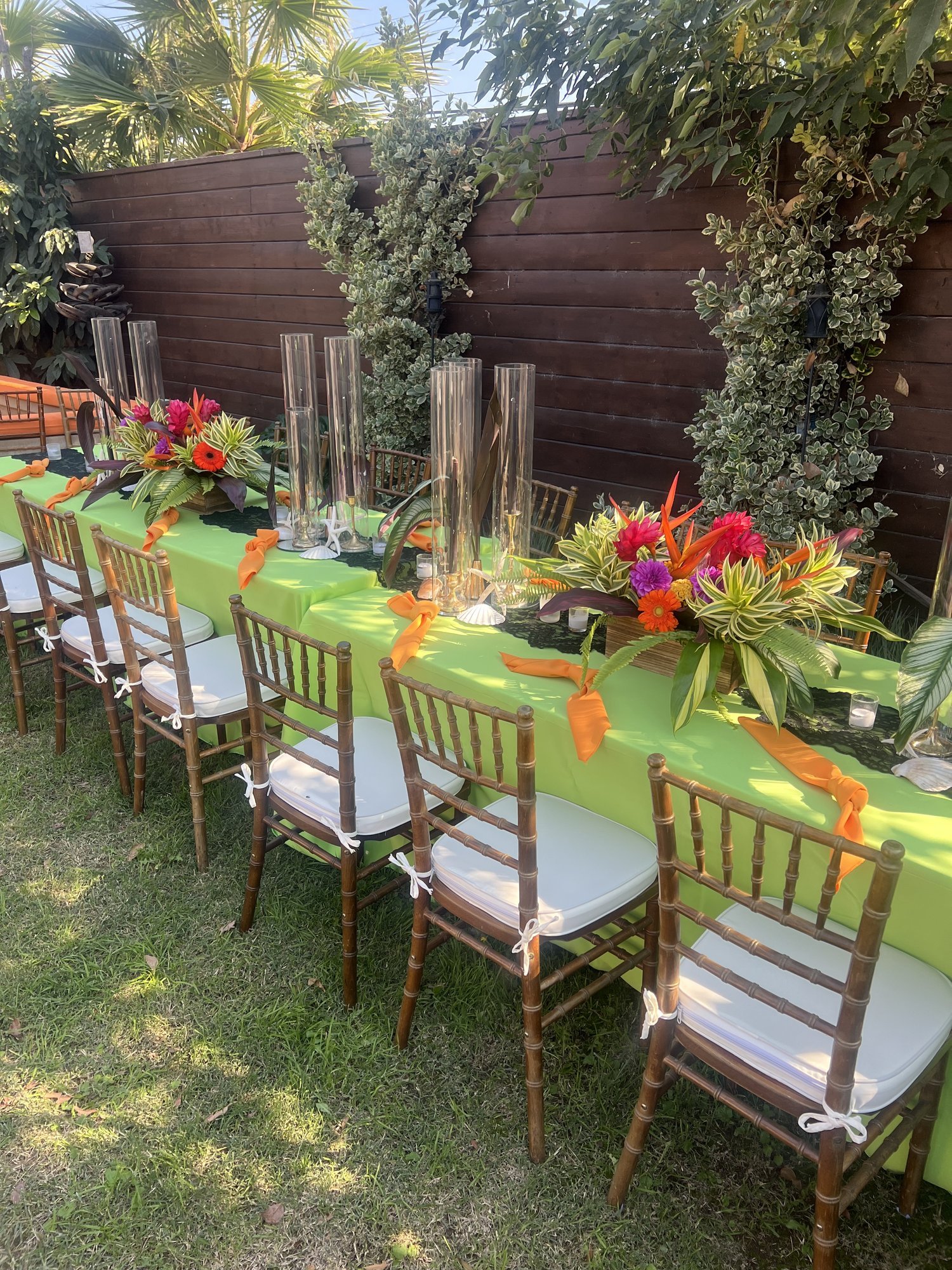 Outdoor table setting with green tablecloth, tall glass vases, colorful tropical flower arrangements, and wooden chairs.