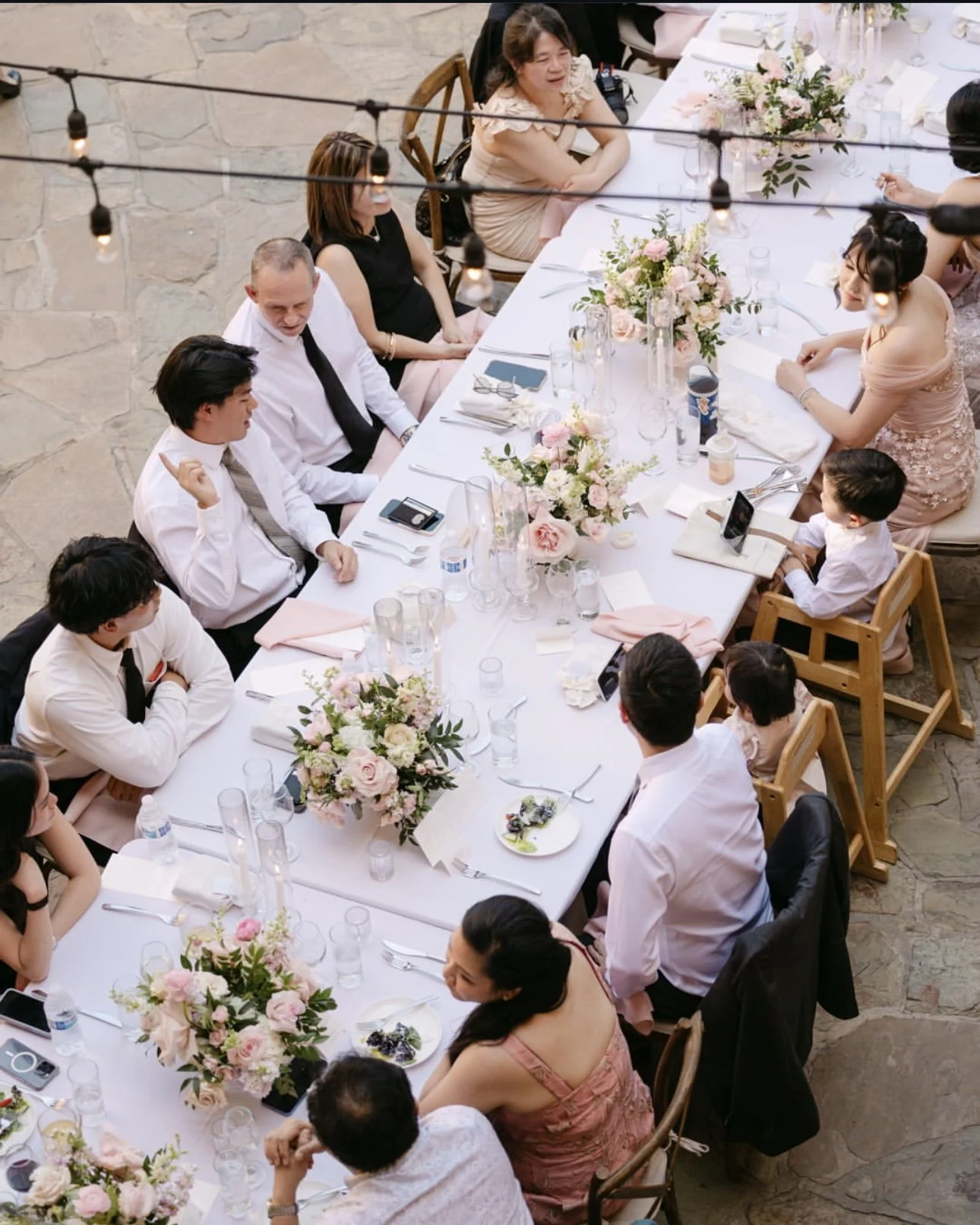 A long dining table decorated with pink and white flower arrangements, set with glassware, plates, and utensils, where people are seated enjoying a formal event or celebration.