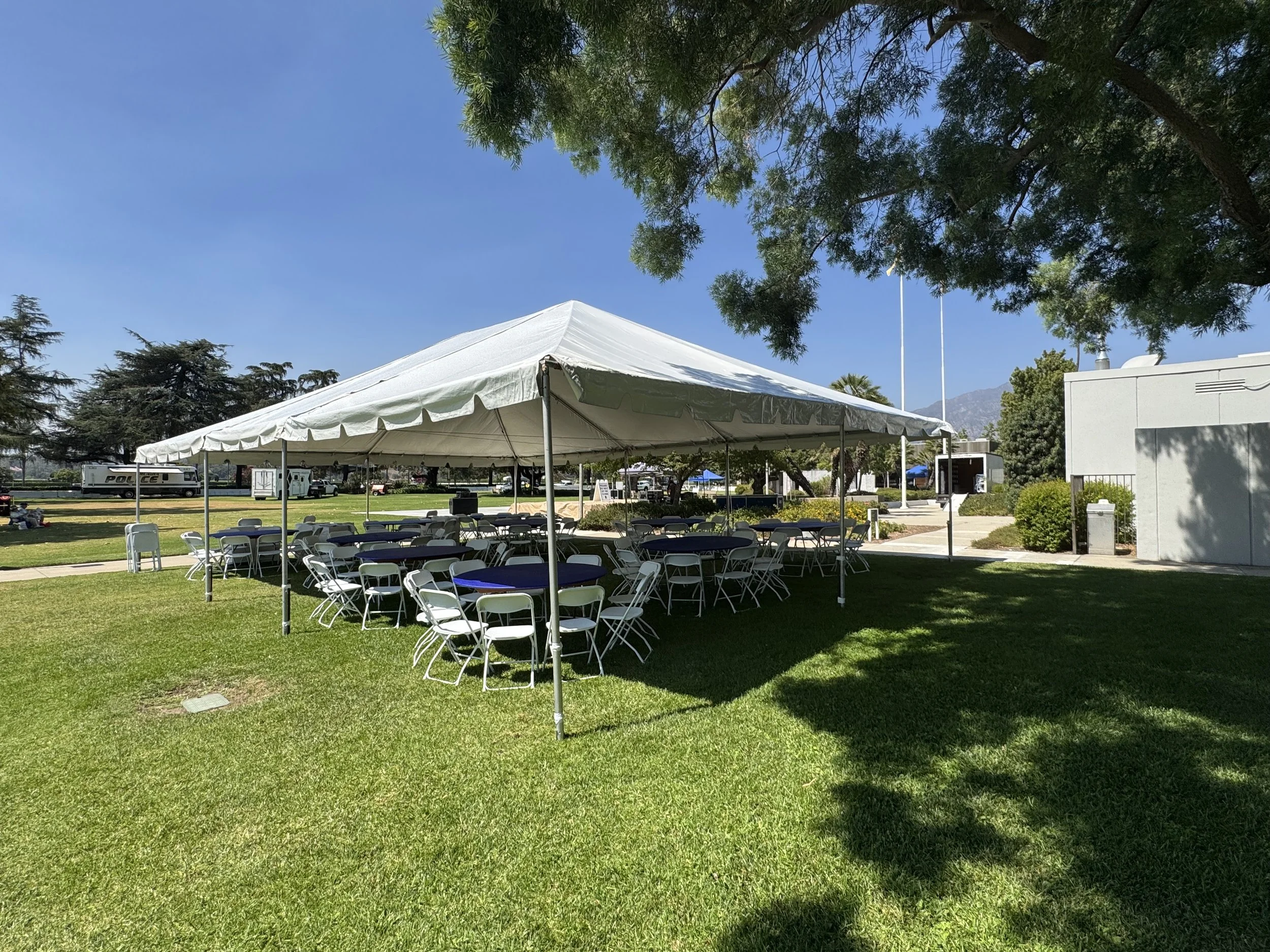 White event tent with multiple tables and chairs set up on a grassy area, with trees and a building in the background, under a clear blue sky.