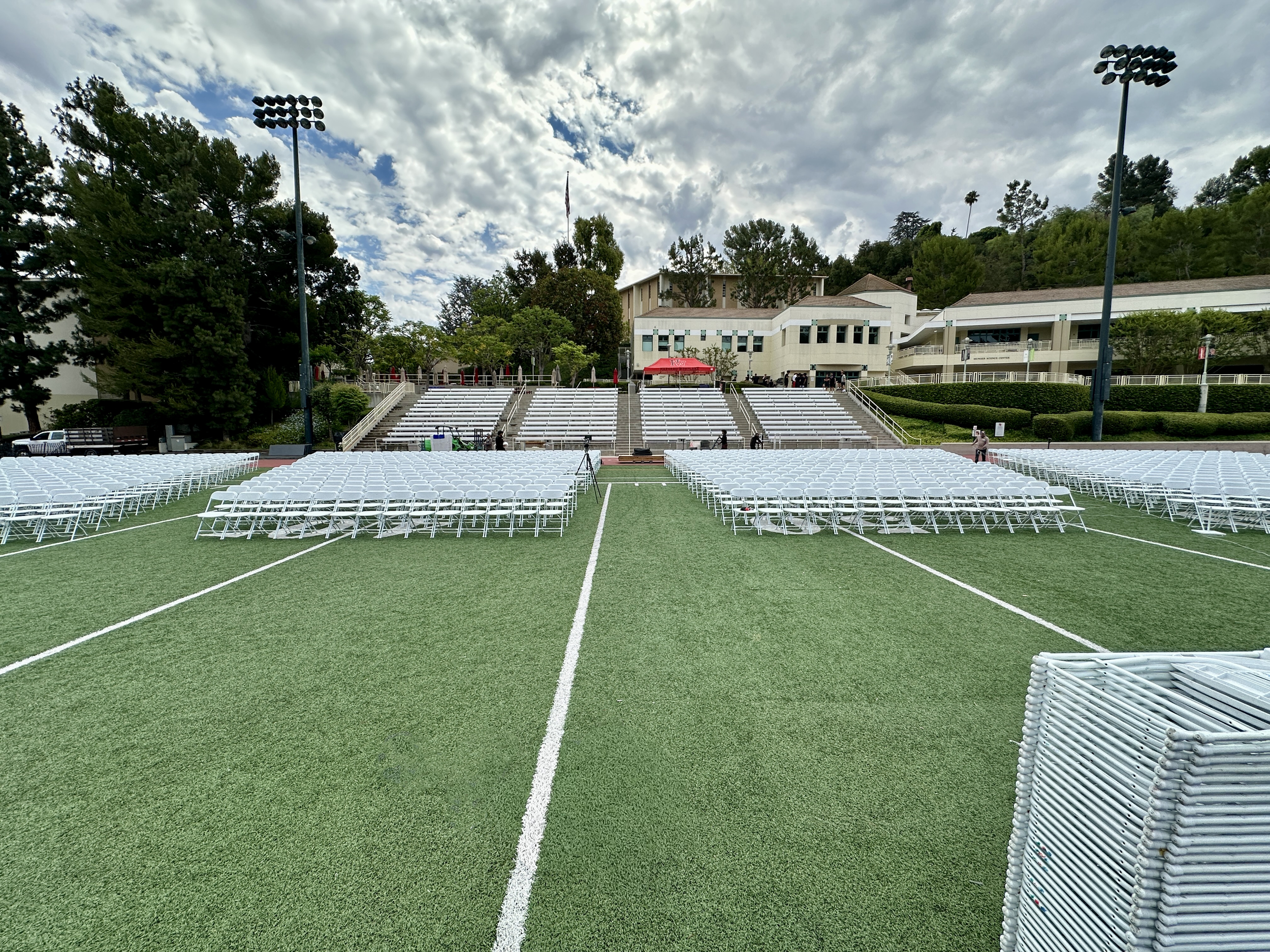 Empty stadium seating with white chairs on a field, prepared for an outdoor event with lighting and buildings in the background, and a cloudy sky overhead.