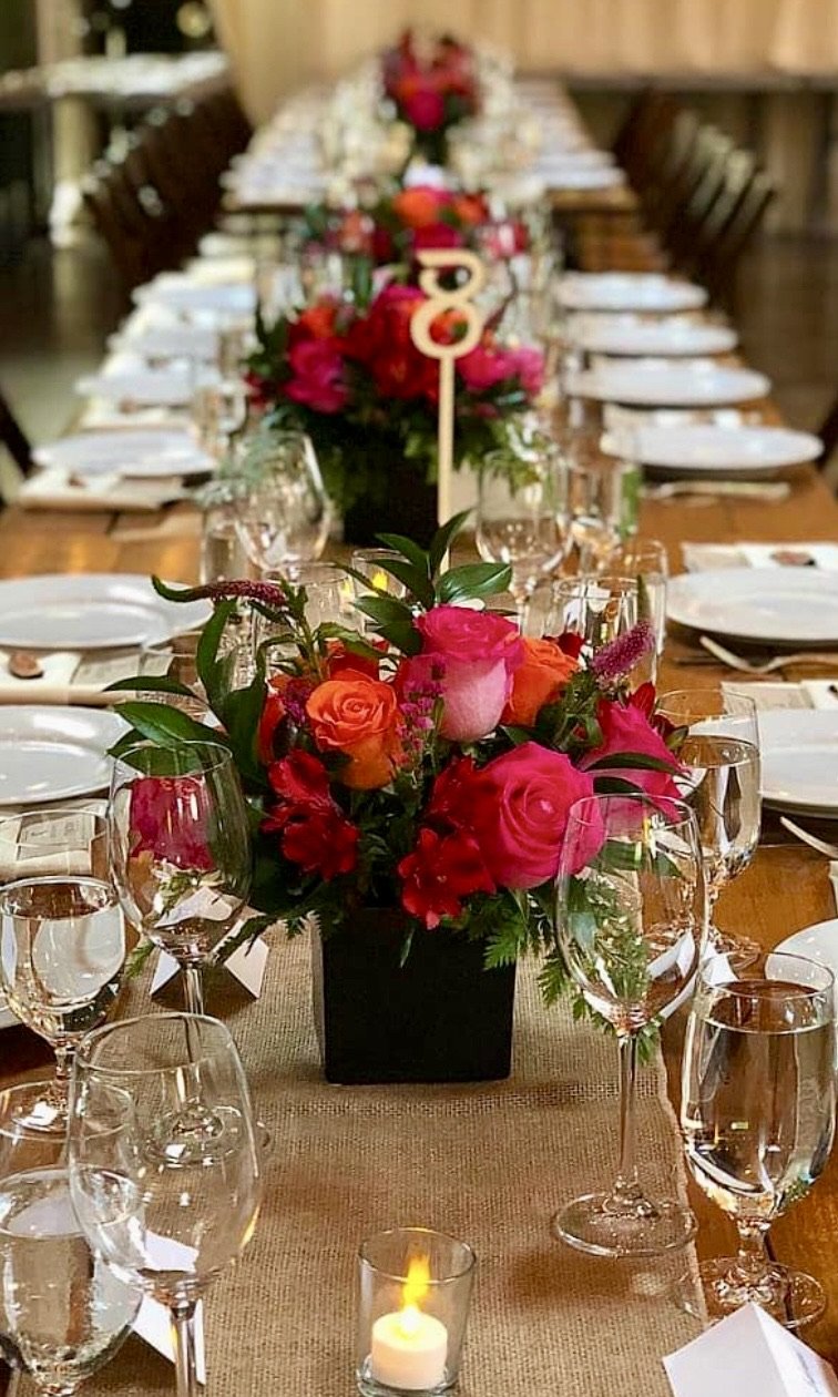 A long dining table decorated with a floral centerpiece featuring pink, orange, and red roses, surrounded by wine glasses, water glasses, and white plates, with a lit candle at the front.