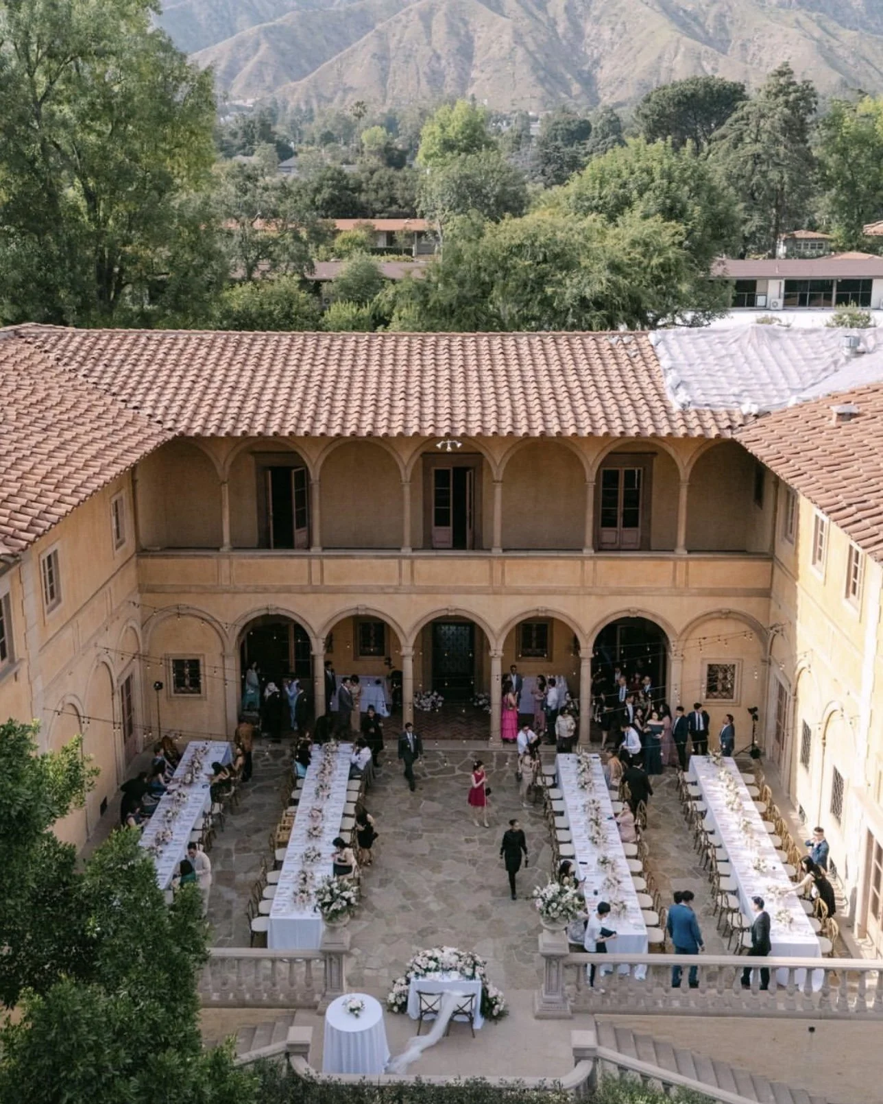 Outdoor wedding reception in a courtyard with long tables, floral arrangements, and wedding guests, surrounded by a historic building with arched walkways and a tile roof, with mountains and trees in the background.