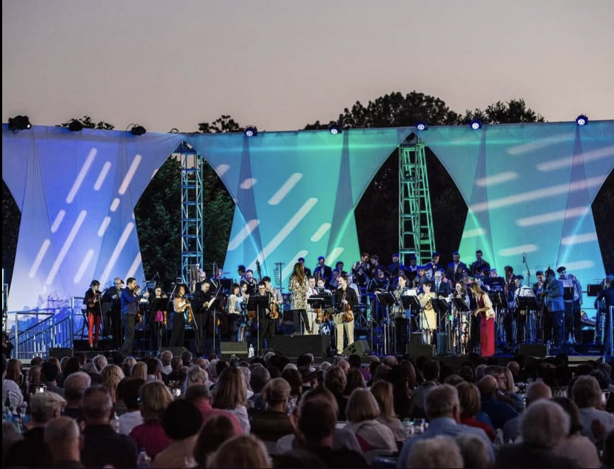 An outdoor orchestra performance on a stage with colorful lighting, surrounded by an audience during dusk.