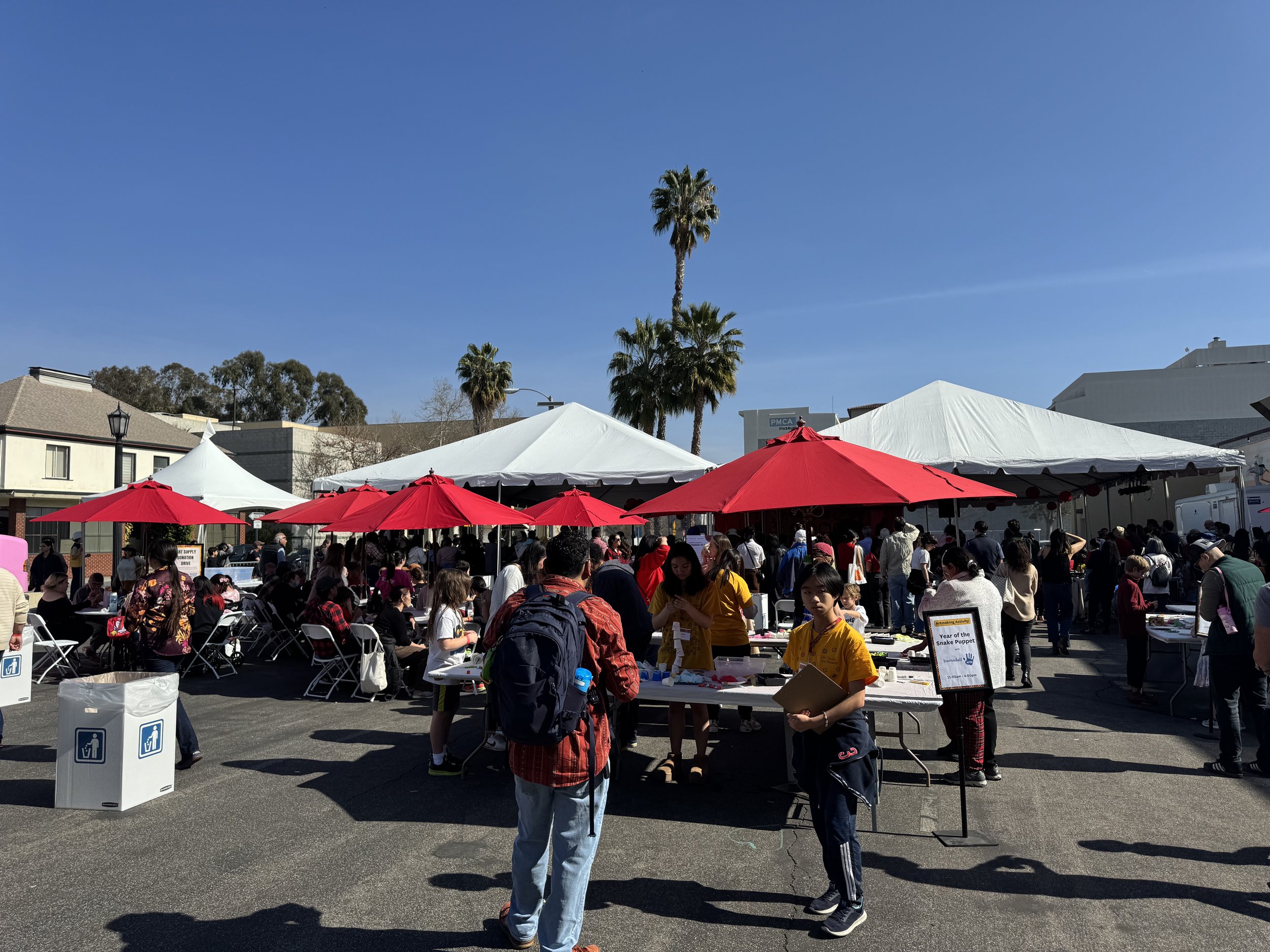 Outdoor event with people gathered under red and white umbrellas, tables with various items, and a clear blue sky.