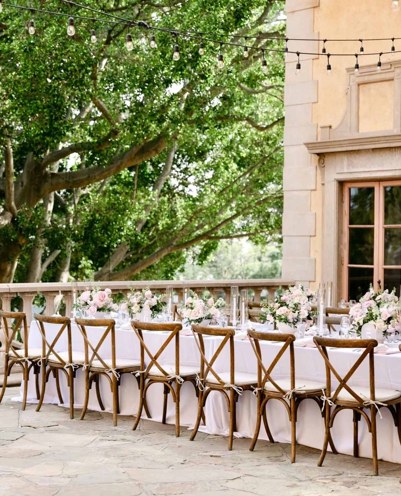 Outdoor banquet table decorated with pink and white flowers, arranged for a celebration or event, under string lights with trees and a stone building in the background.