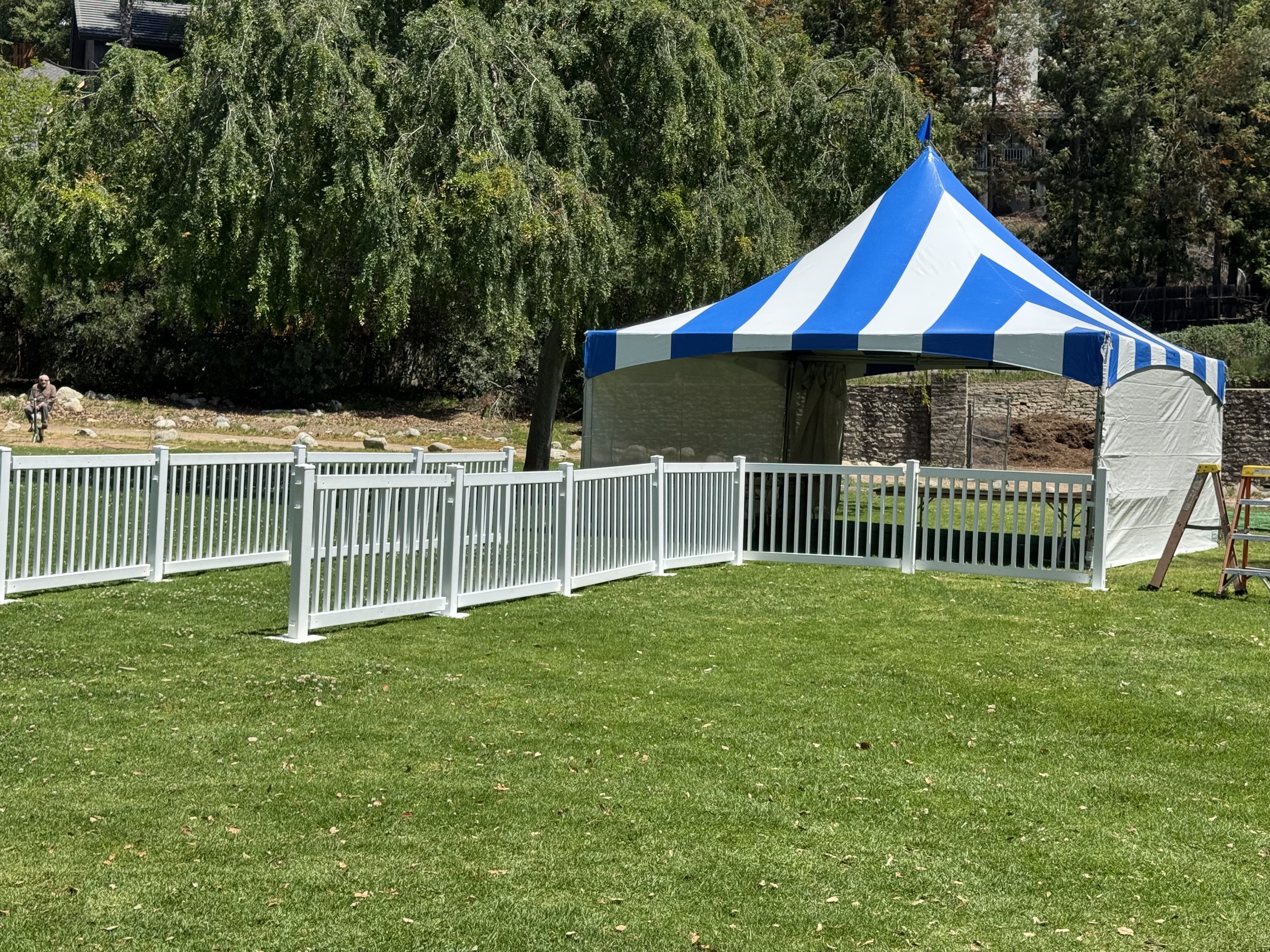 A blue and white striped canopy tent set up on a grassy area, surrounded by a white picket fence. There is a ladder nearby and trees in the background.