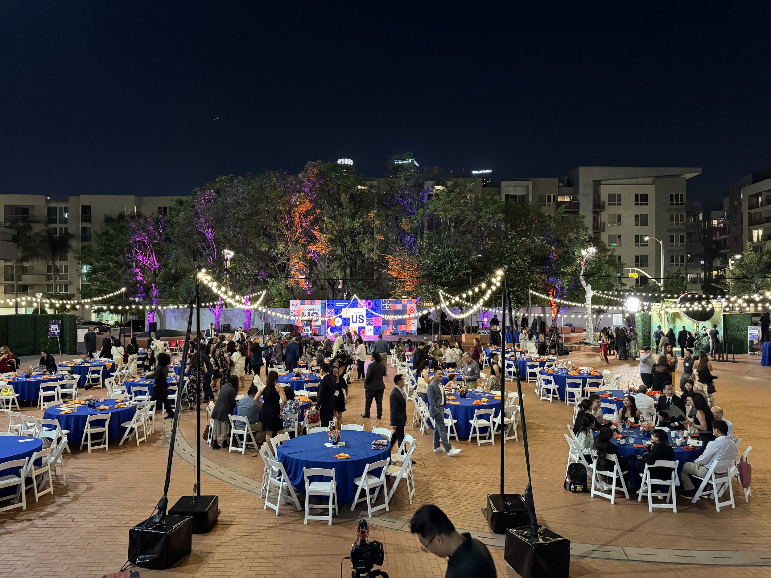 Outdoor event with tables and chairs set up on a large patio or plaza, featuring string lights, a stage or display area, and people mingling and sitting at the tables.