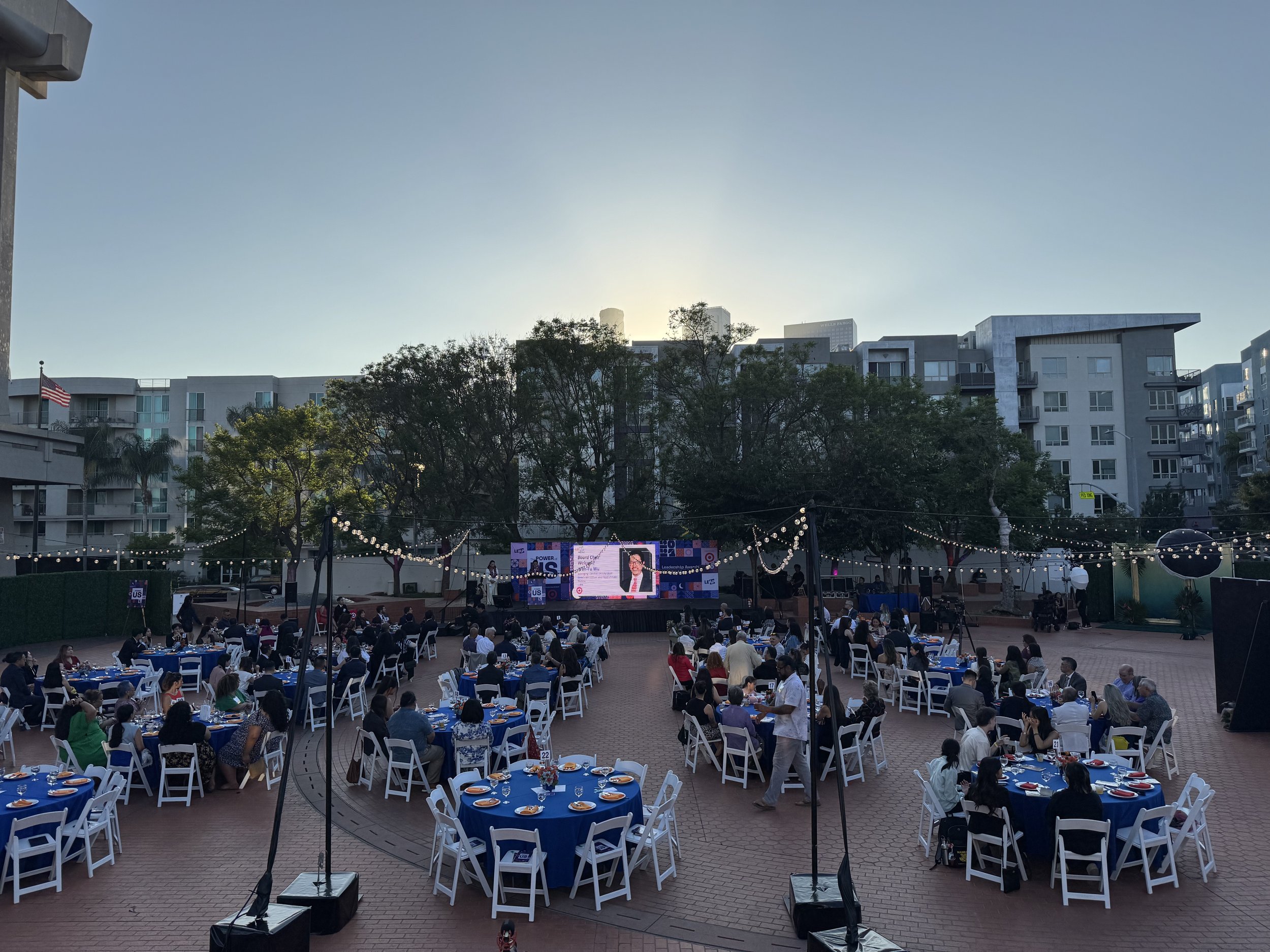 Outdoor event with tables covered in blue tablecloths and white chairs, attended by people sitting and socializing. A stage with a screen displays a presentation, surrounded by string lights. Buildings and trees form the background, with an American 