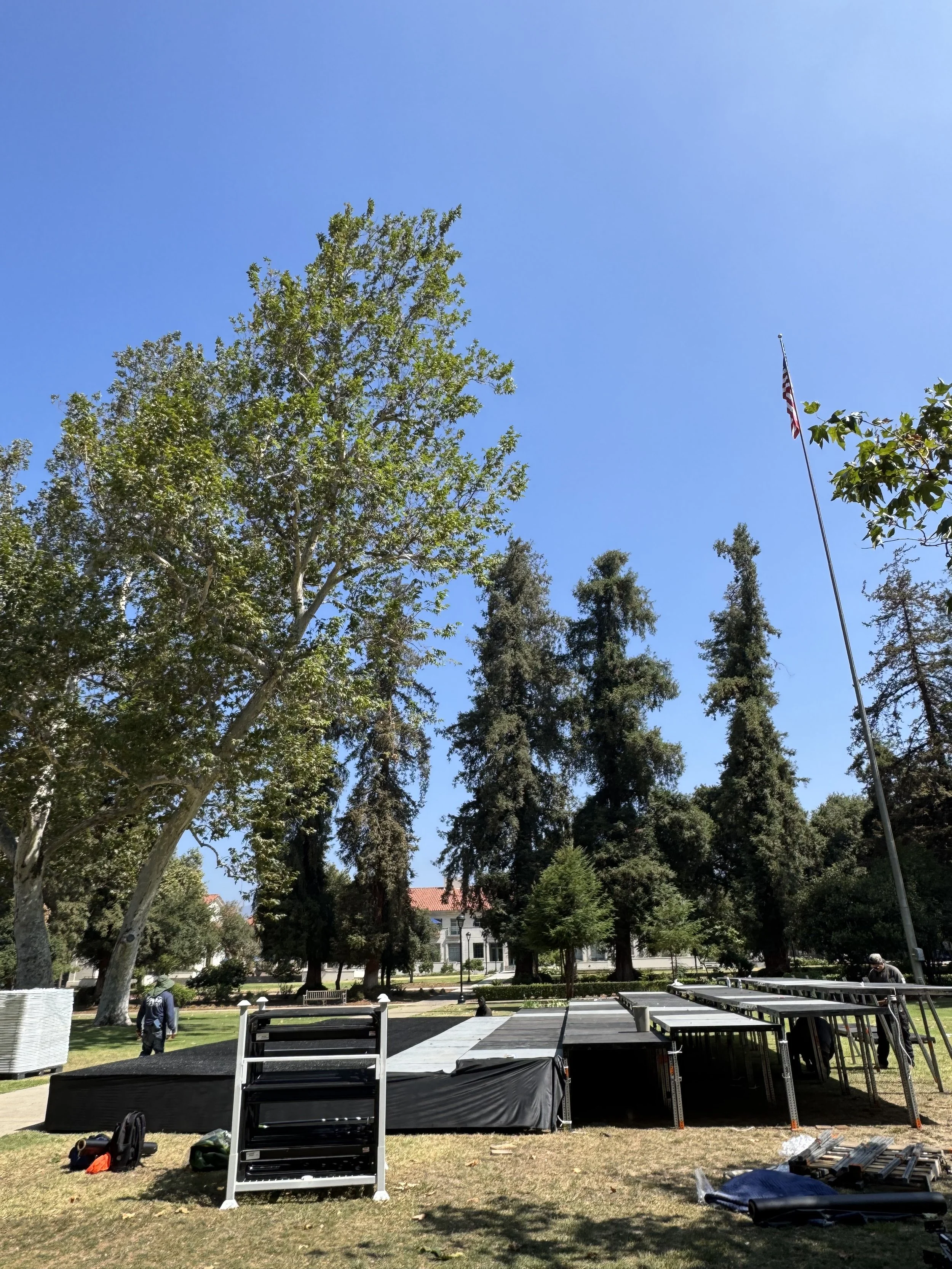 Setting up outdoor stage with metal scaffolding and stairs, trees and American flag in park or public area on a sunny day.