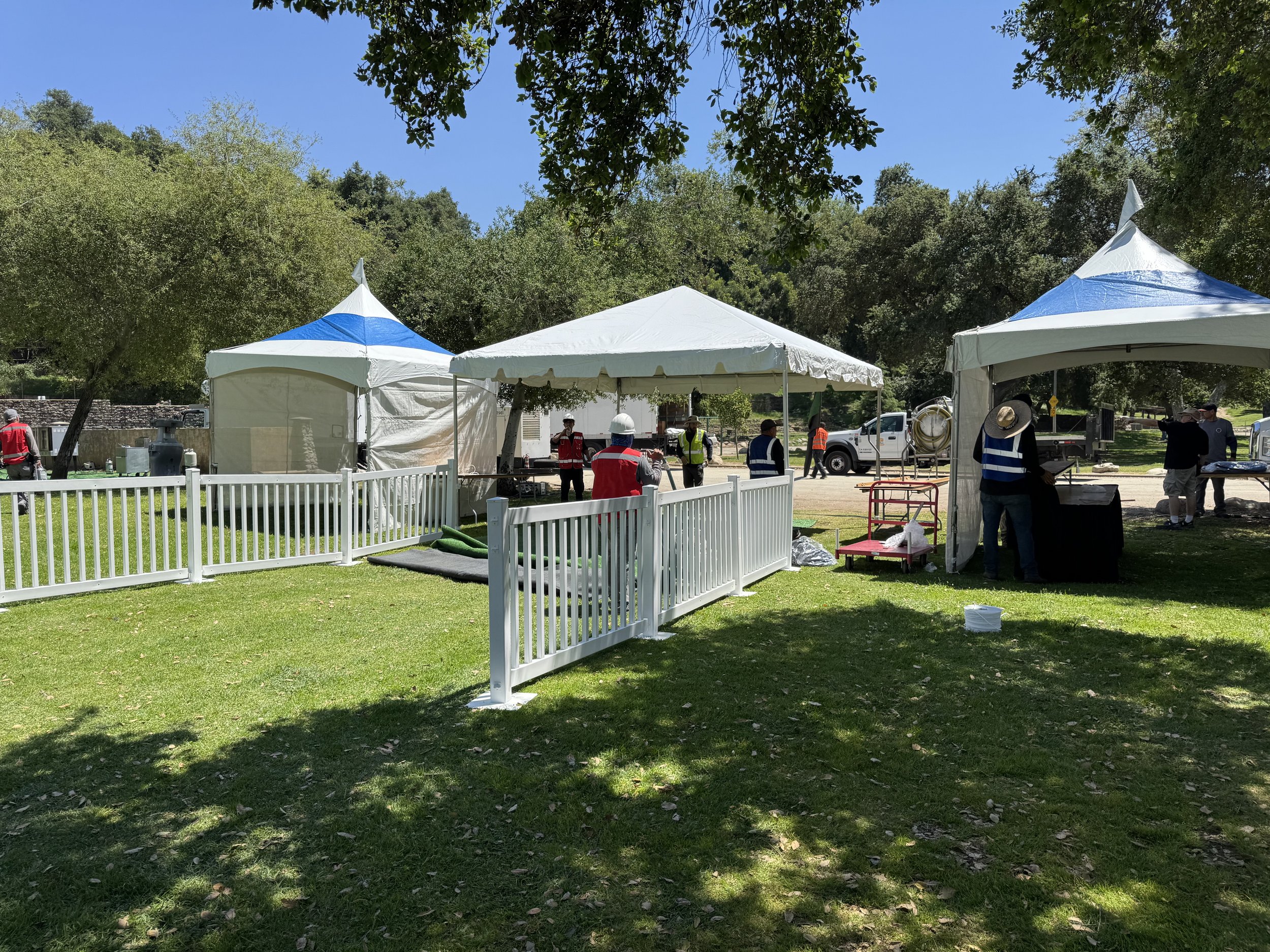 Outdoor event setup with white tents, blue canopies, white fencing, and people in safety vests on a grassy area.