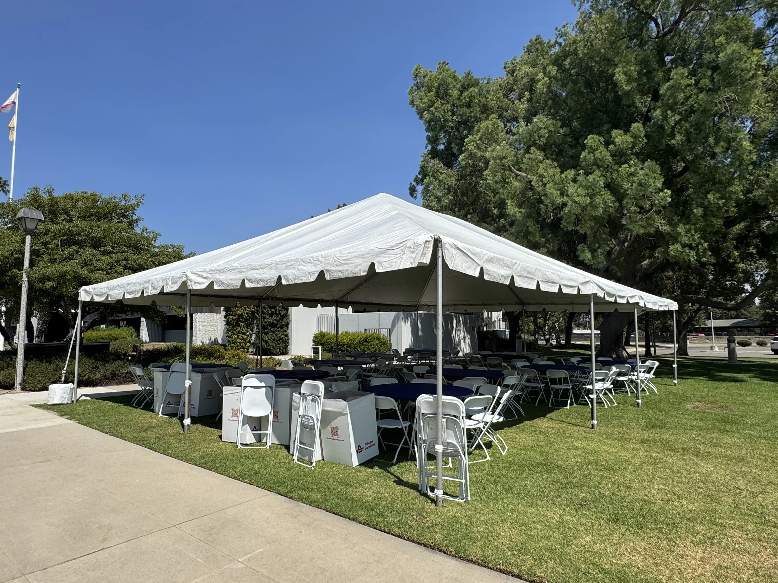 White event tent set up on a grassy area with folding chairs and tables underneath. Trees and a clear blue sky in the background.