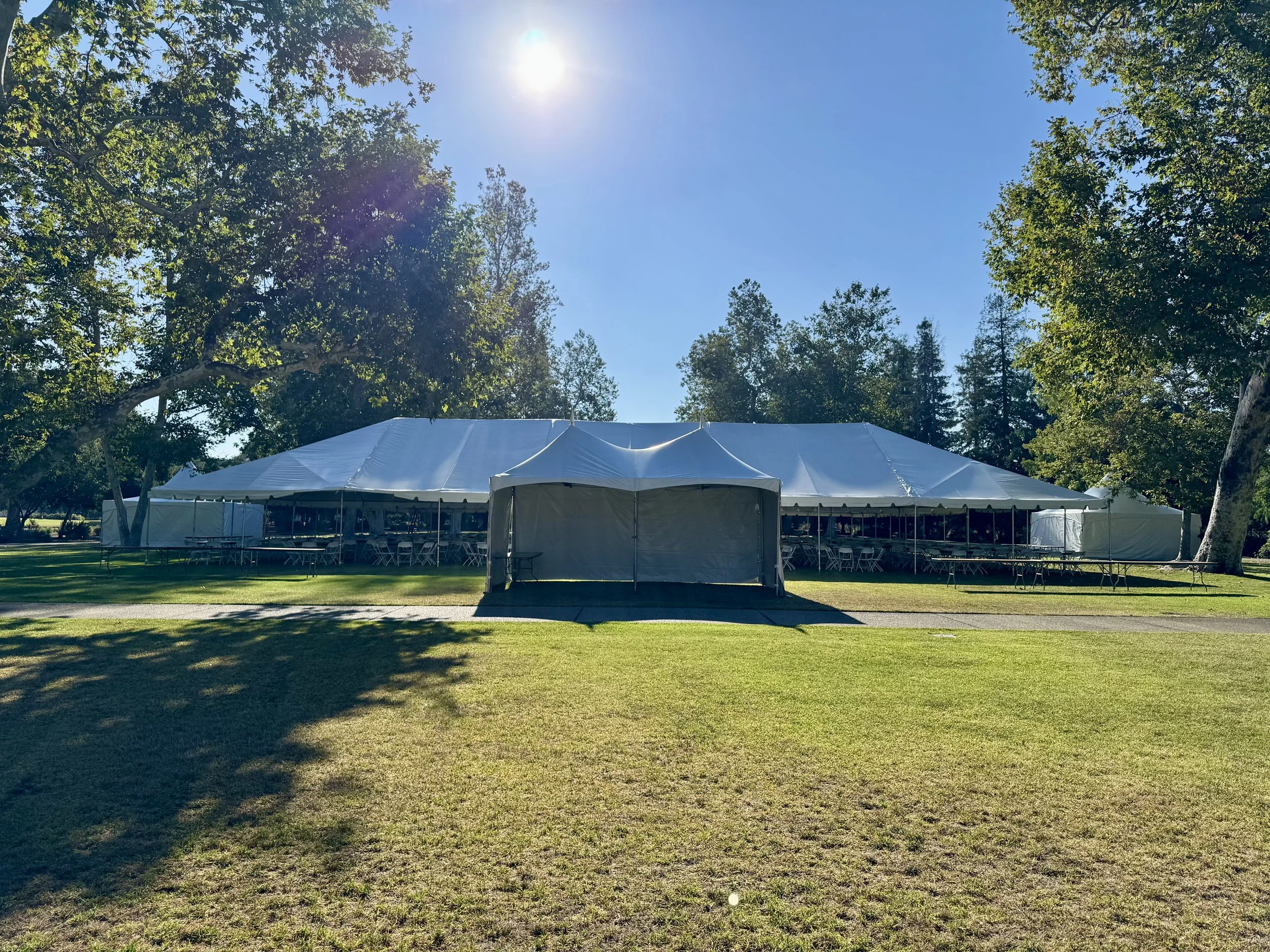 Large white event tent set up on a grassy outdoor area surrounded by trees, with tables and chairs inside, under a clear blue sky with the sun shining brightly.