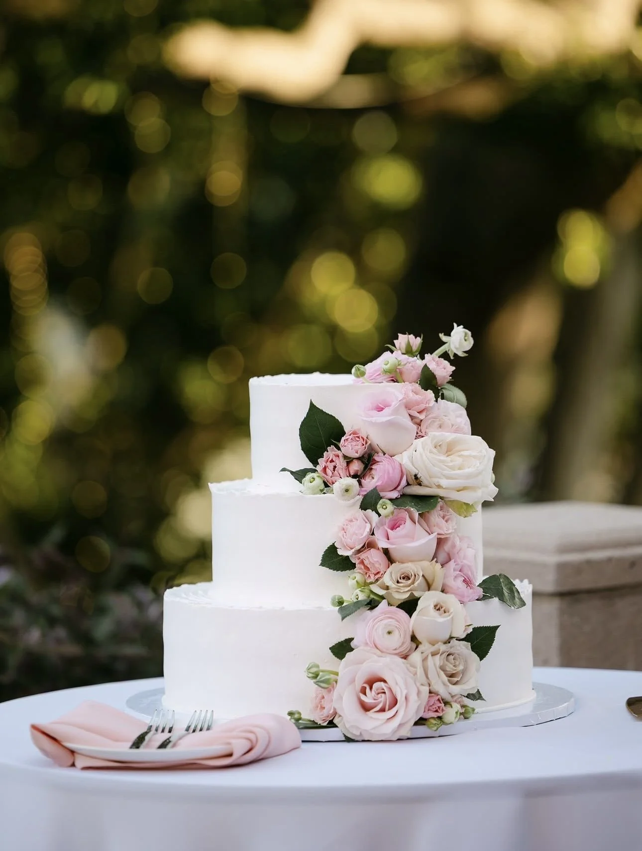 A three-tier white wedding cake decorated with cascading pink, white, and cream roses and greenery, placed on a white table with a pink napkin and silver forks.