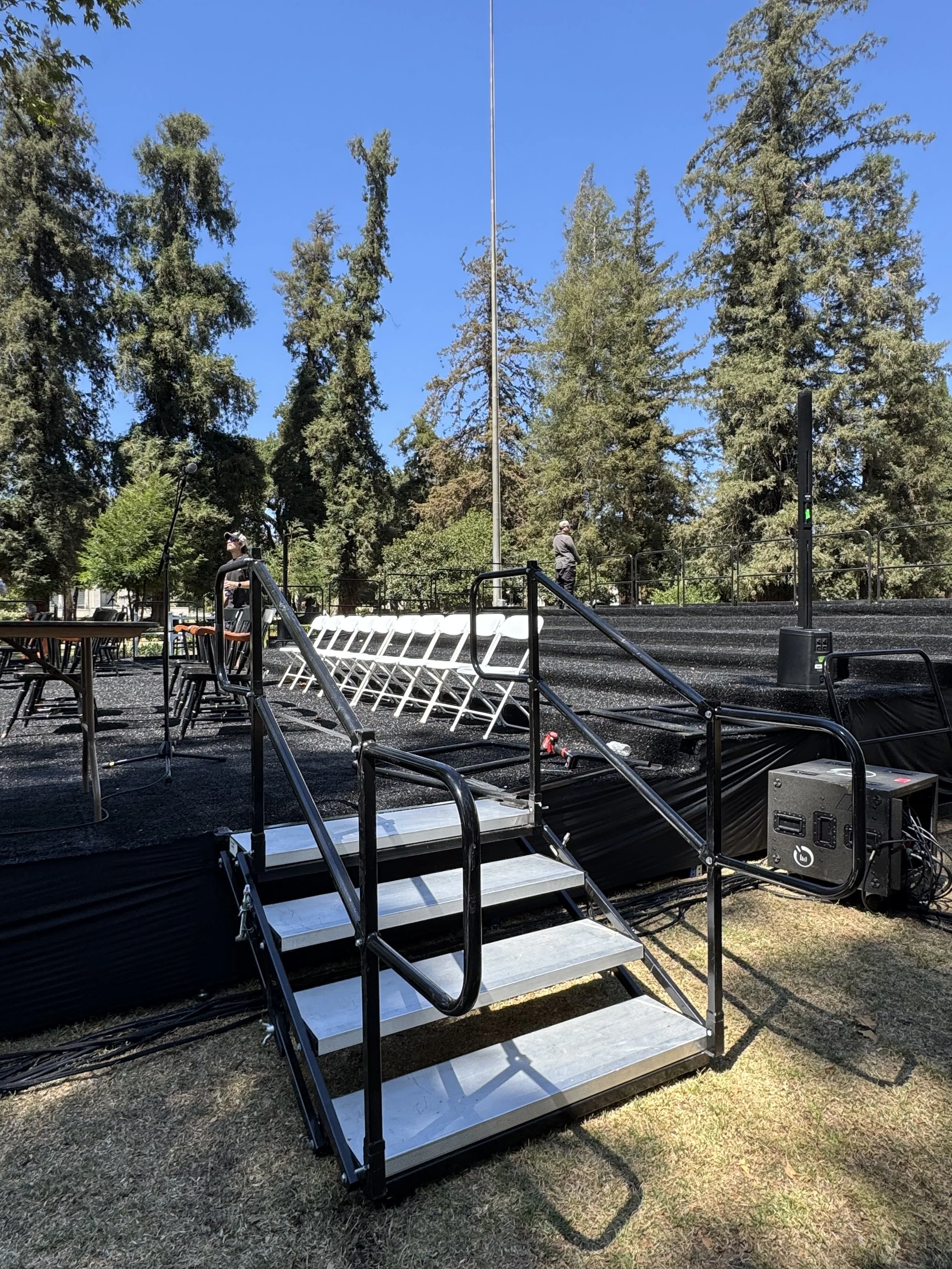 Outdoor stage with metal stairs, black railing, and audio equipment, set in a park with tall trees and a clear blue sky.