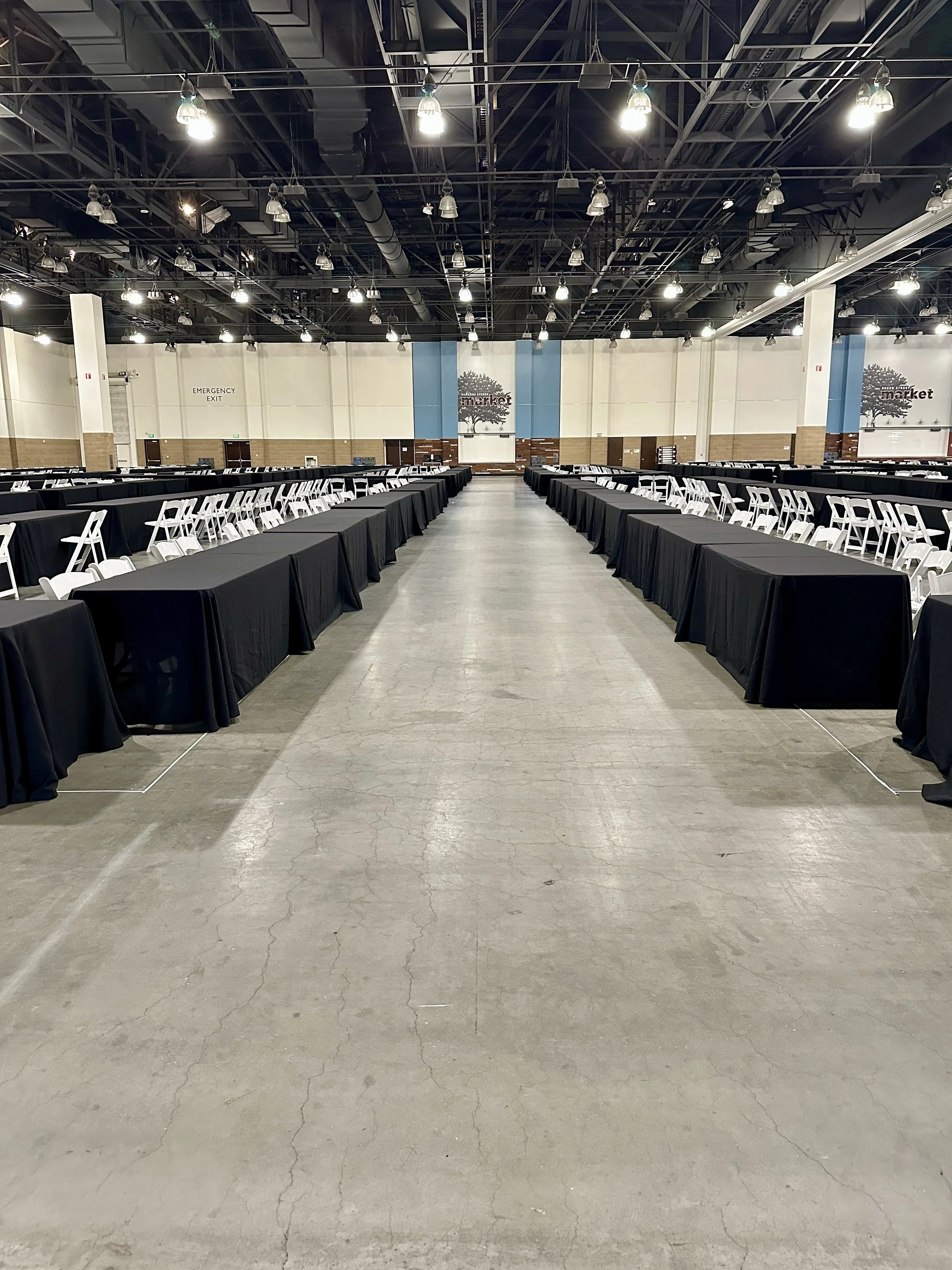 Large conference hall with long black-tabled setups and white chairs, overhead lighting, and two signs indicating emergency exits.