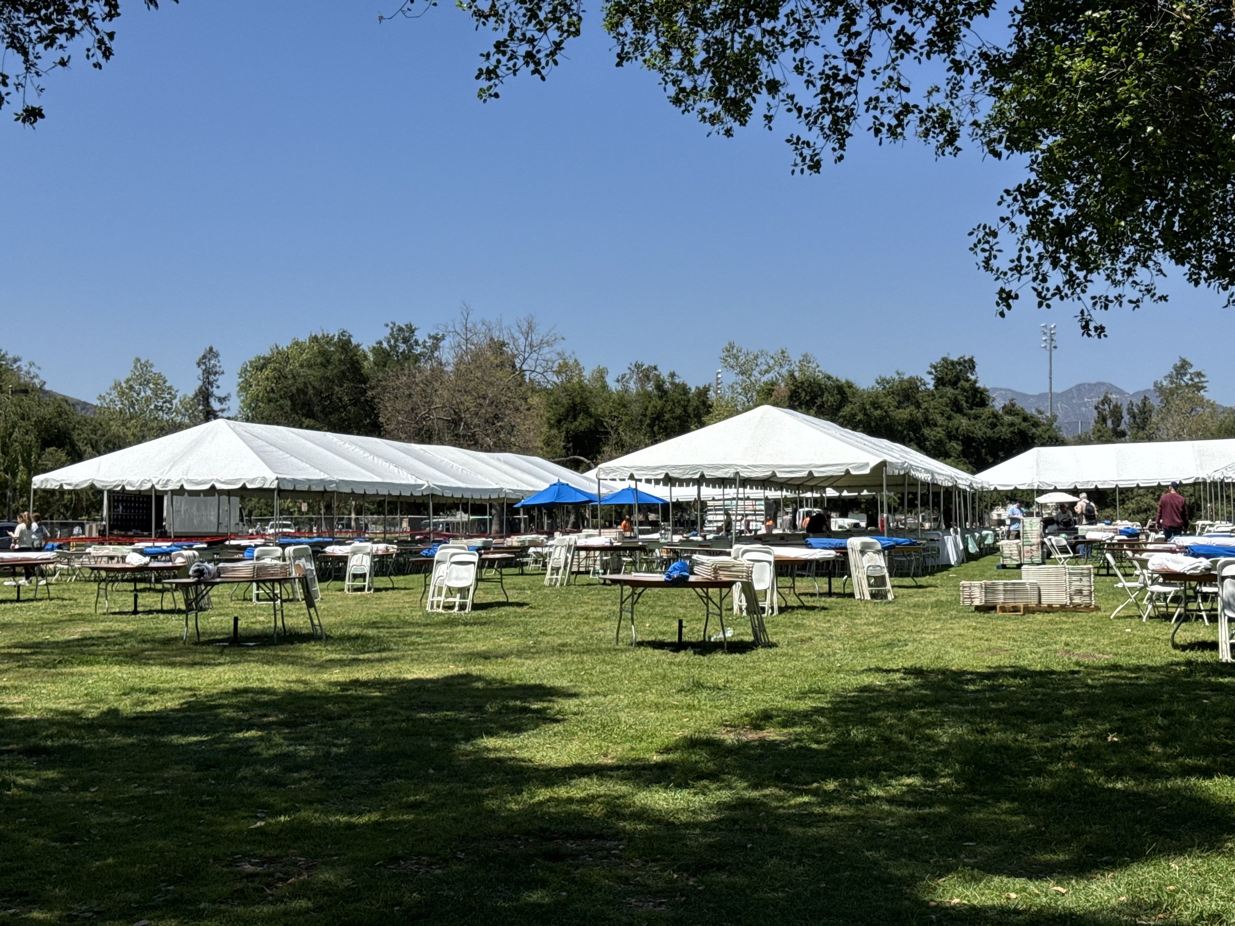Outdoor event setup with large white tents, tables, and chairs on a grassy field under clear skies.