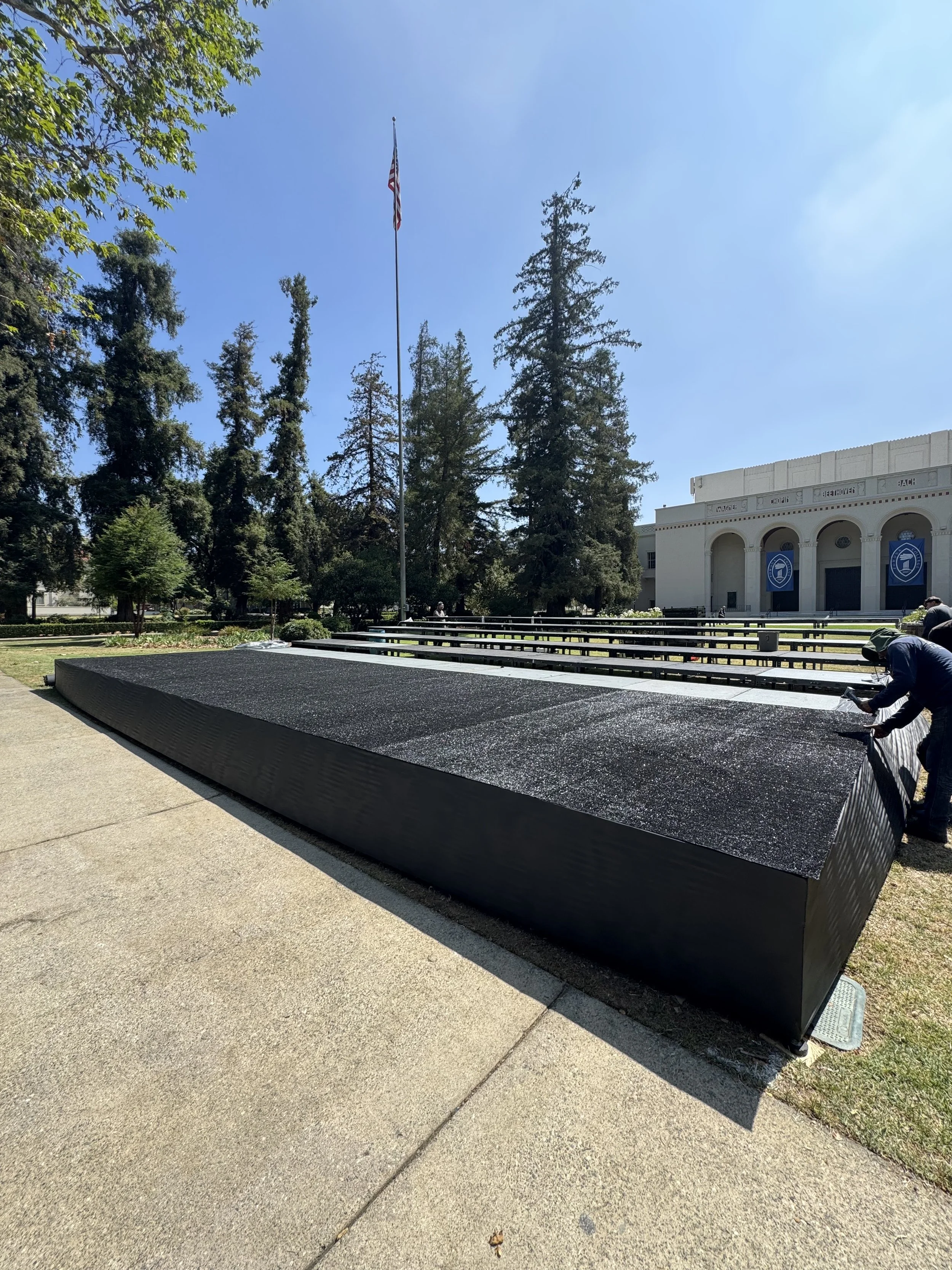 People installing a black granite memorial with the American flag flying on a flagpole nearby, in front of a white building with arches, outdoor area with benches, trees, and a blue sky.