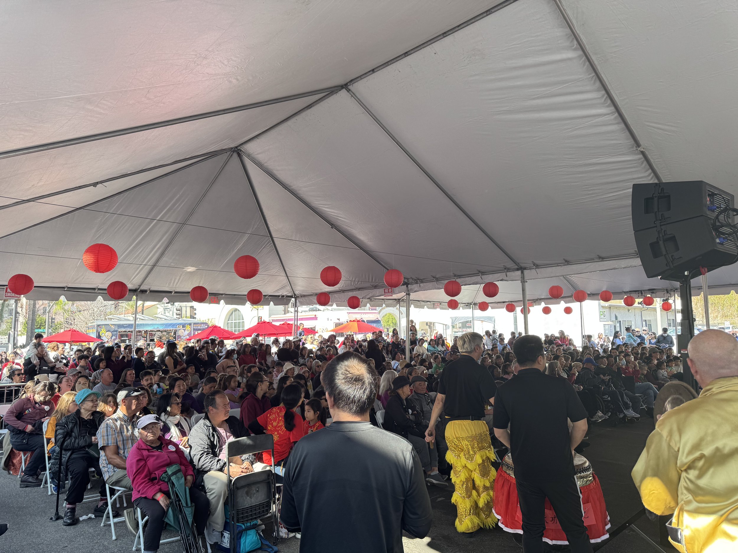 Crowded event under a large tent with red paper lanterns and people seated and standing, some in traditional attire, at a cultural festival.