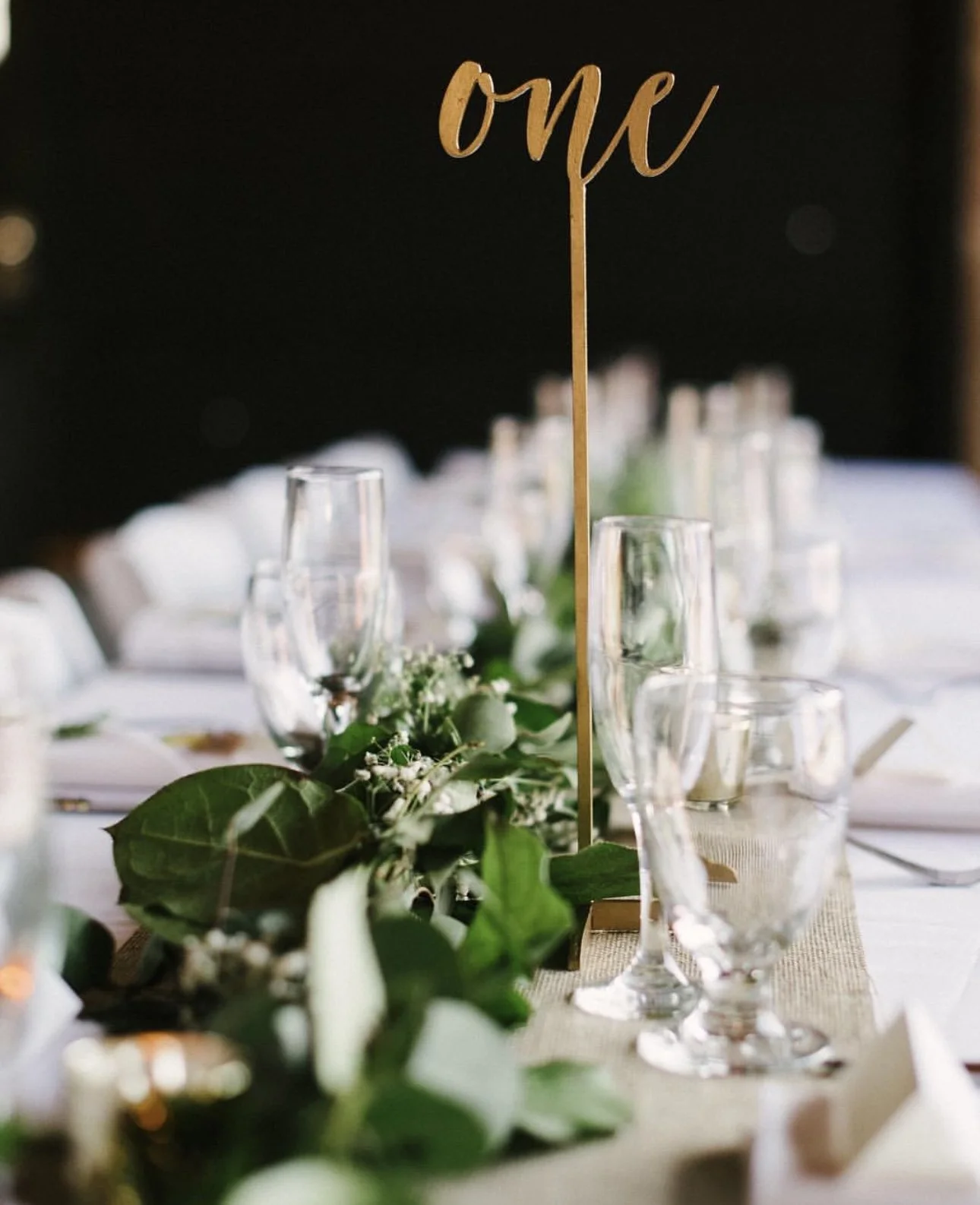Table setting with glassware and greenery centerpiece, and a gold table number sign that says 'one' for a celebration or wedding.