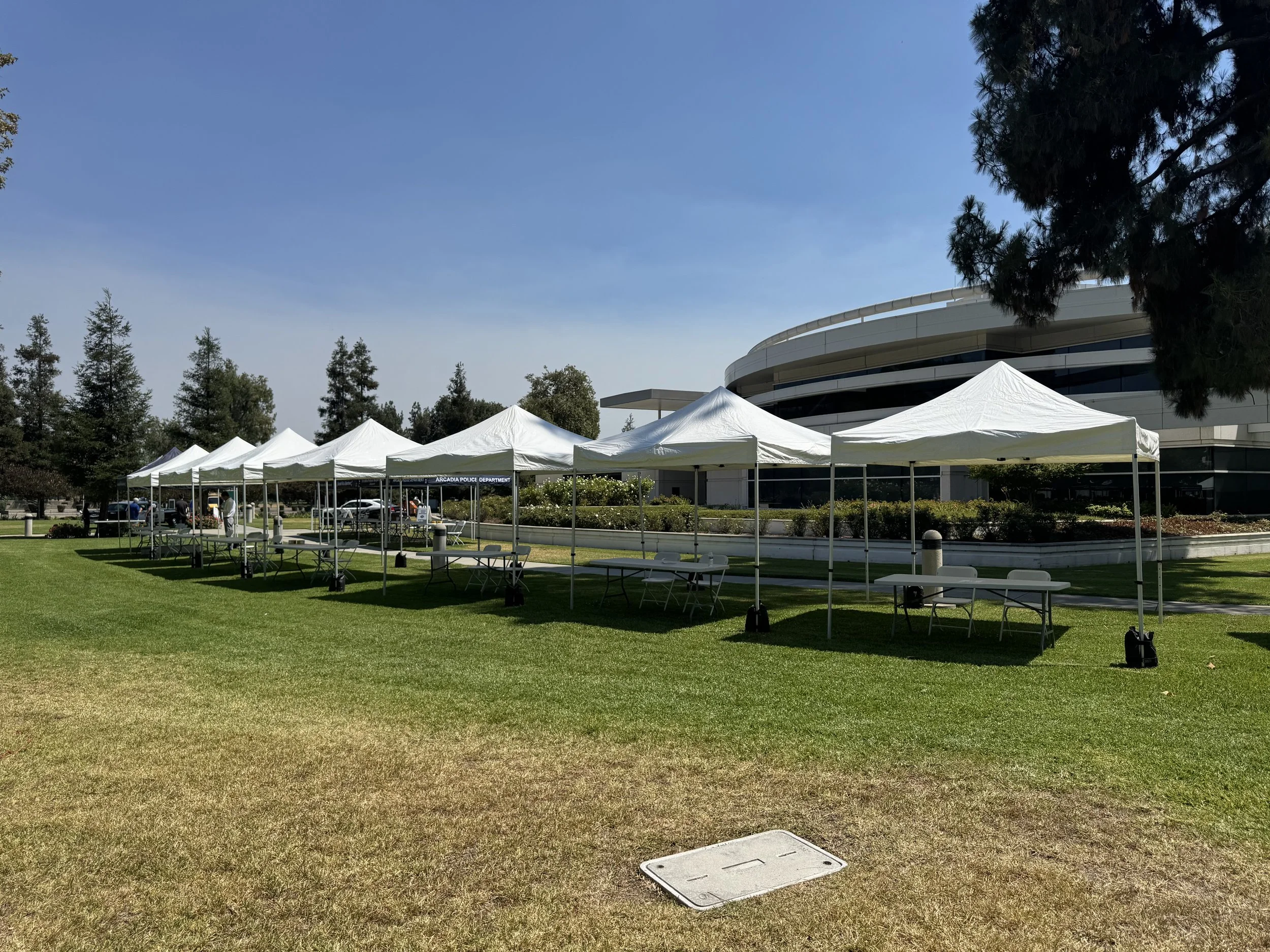 Multiple white tents set up on a grassy lawn with a large modern building in the background, trees surrounding the area, and a clear blue sky.