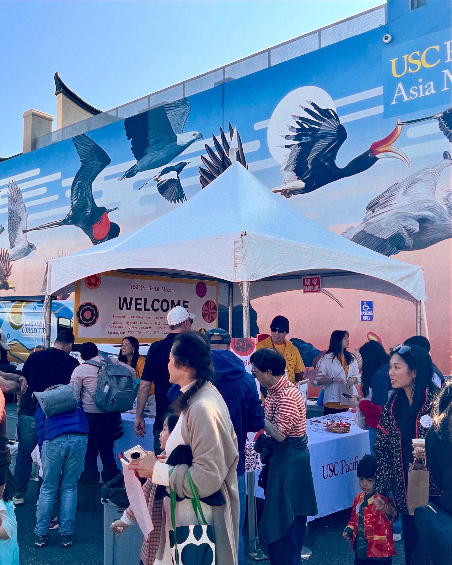 Outdoor event at USC Pacific Asia Museum with a mural of birds, a welcome tent, and people gathering.