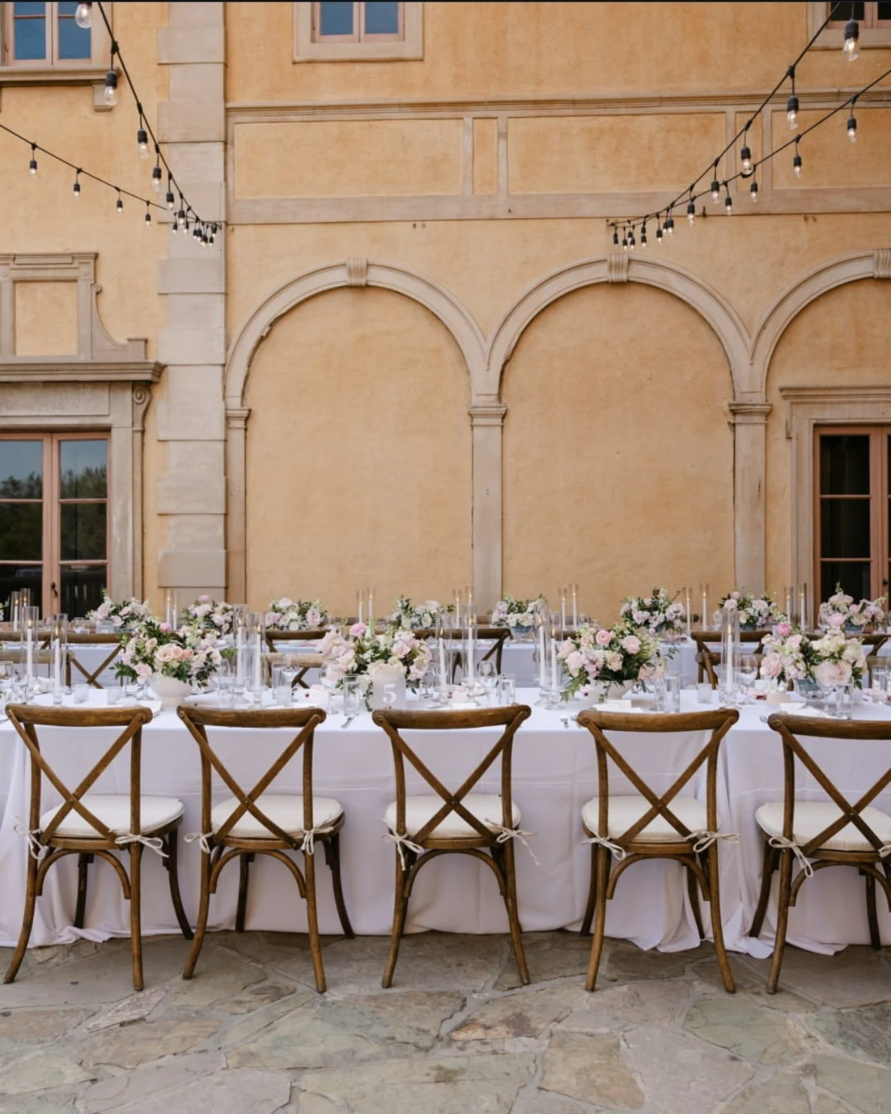 Elegant outdoor reception table set with pink and white floral arrangements, glassware, candles, and wooden chairs with tied ribbons, under string lights, against a yellow stucco building.