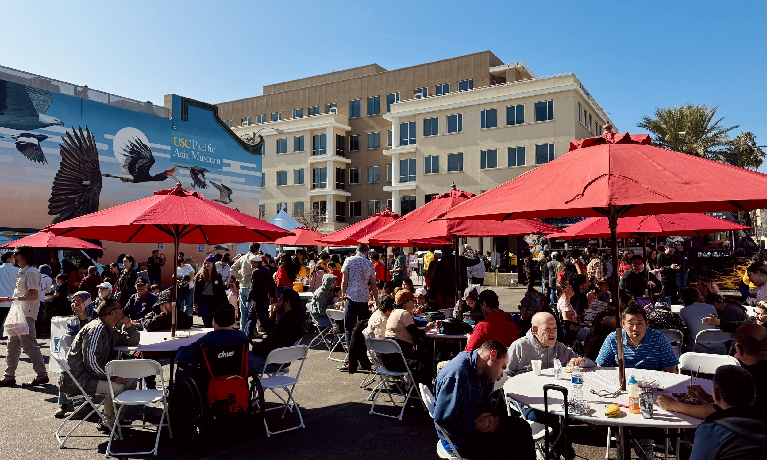 Outdoor event with red umbrellas, people sitting at tables, mural of birds on building, USC Pacific Asia Museum sign.