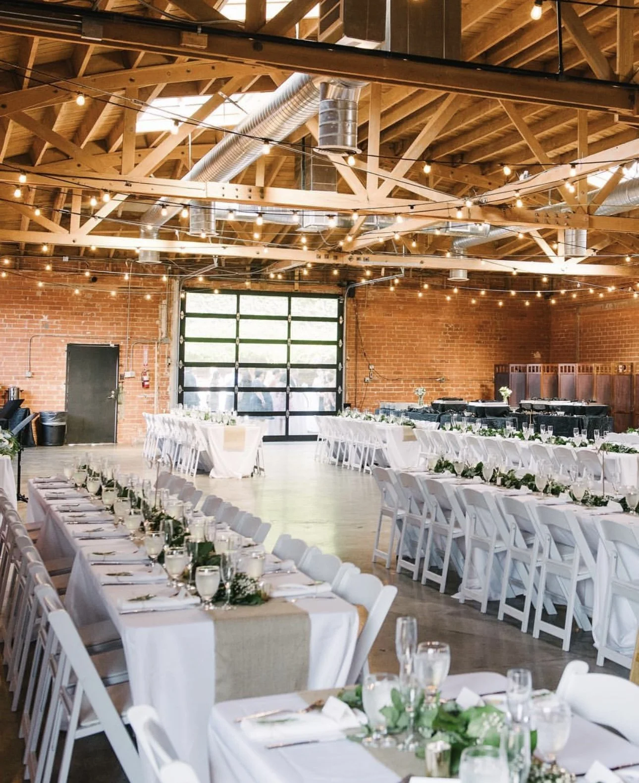 Interior of a wedding or event reception hall with long rectangular tables decorated with white tablecloths, greenery, and candles. The room has exposed brick walls, large garage-style door, and string lights hanging from the wooden ceiling beams.