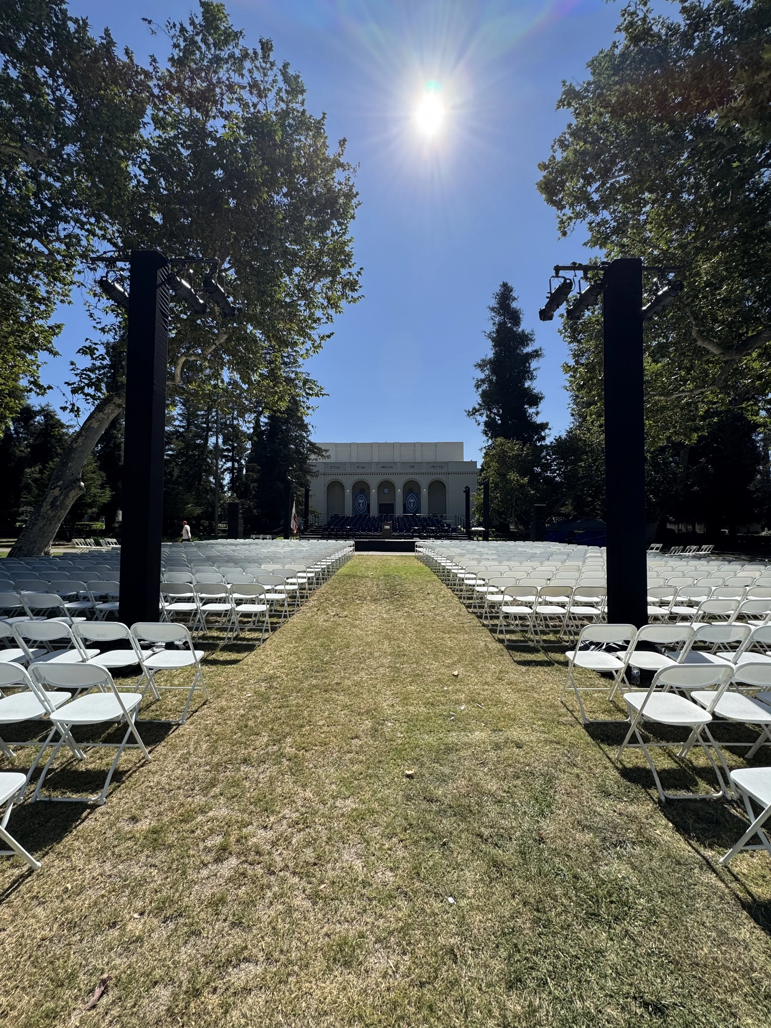 Outdoor event setup with rows of white chairs facing a stage under a building, with tall trees and bright sun overhead.