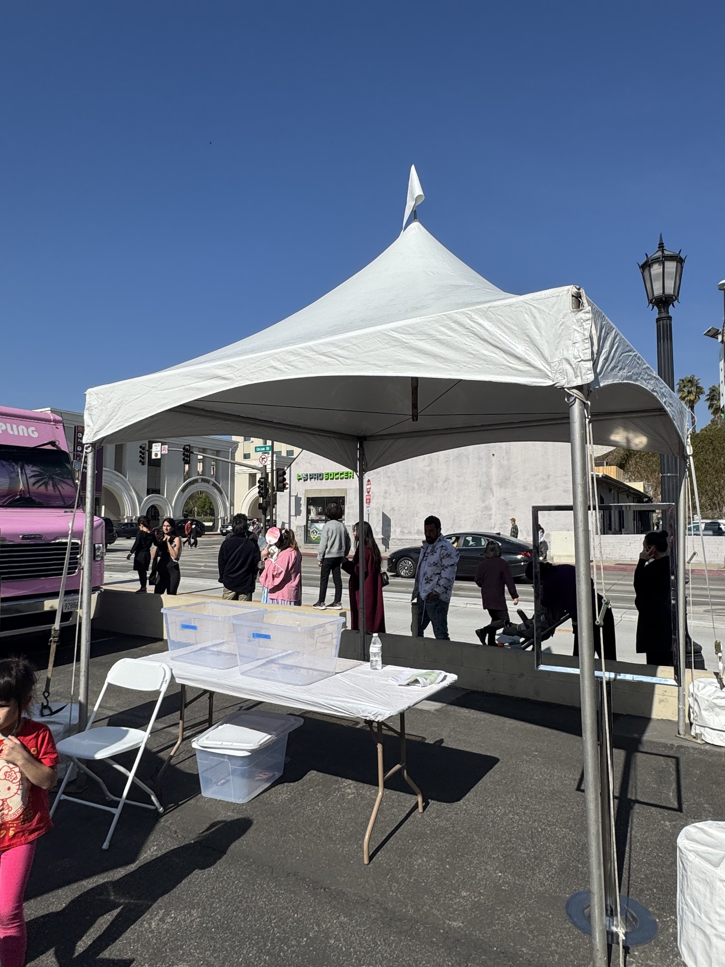 Outdoor tent setup with a white canopy and metal frame, featuring a table with empty clear bins, a folding chair, and a water bottle. People are walking around in the background, and a pink vehicle is partially visible on the left. It's a sunny day w