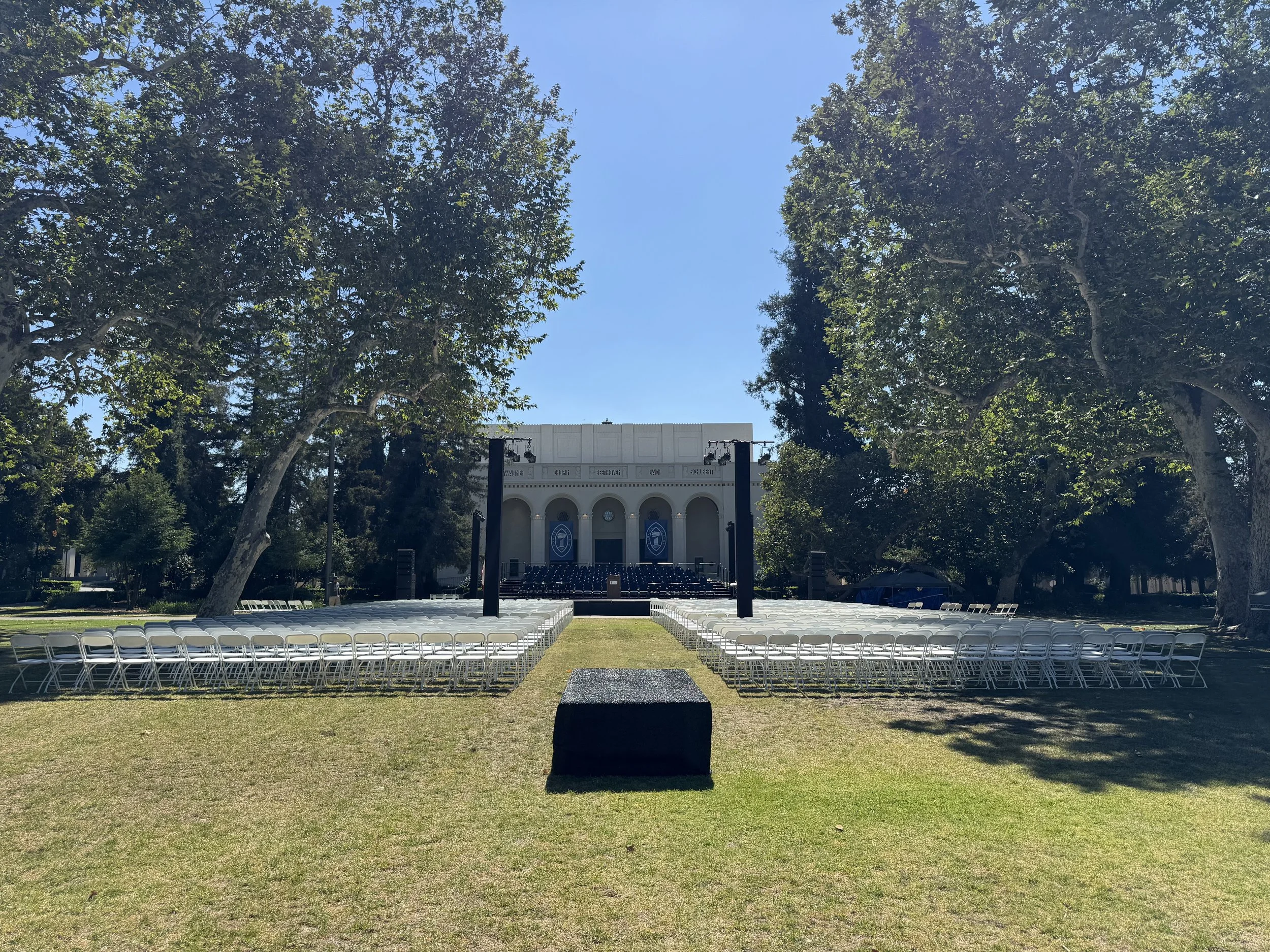 Outdoor stage setup in a park with white chairs arranged on both sides, large trees, and a white building with arches in the background.