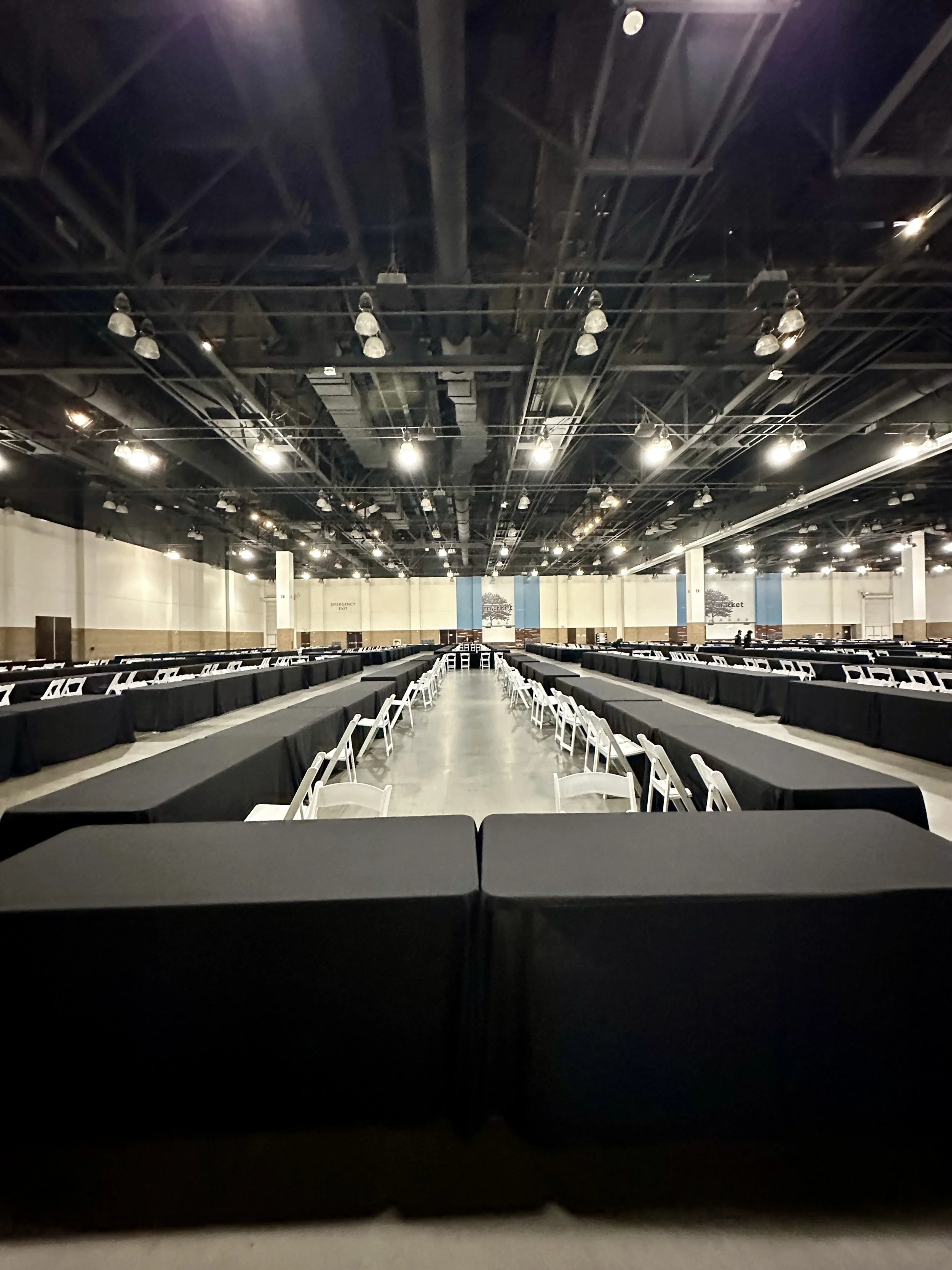 Empty conference hall with long black tables, white chairs, and a high ceiling with exposed pipes and hanging lights.