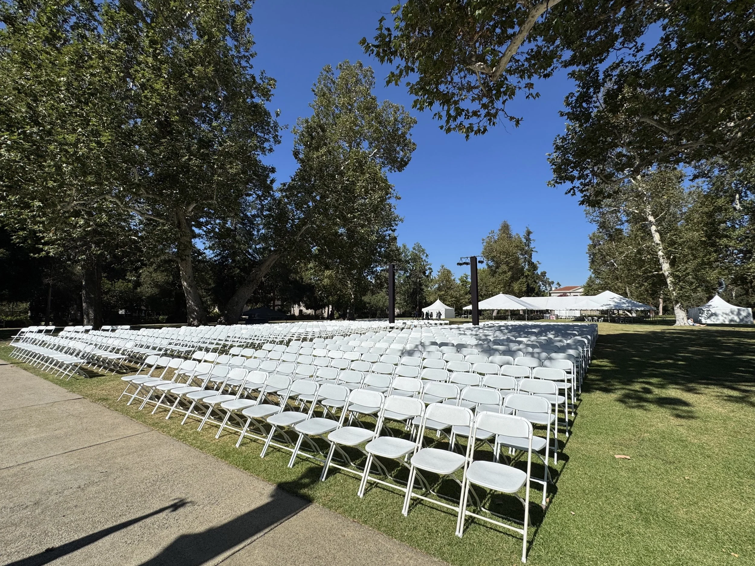 Empty white chairs arranged in rows for an outdoor event on a grassy area under large trees with tents in the background.