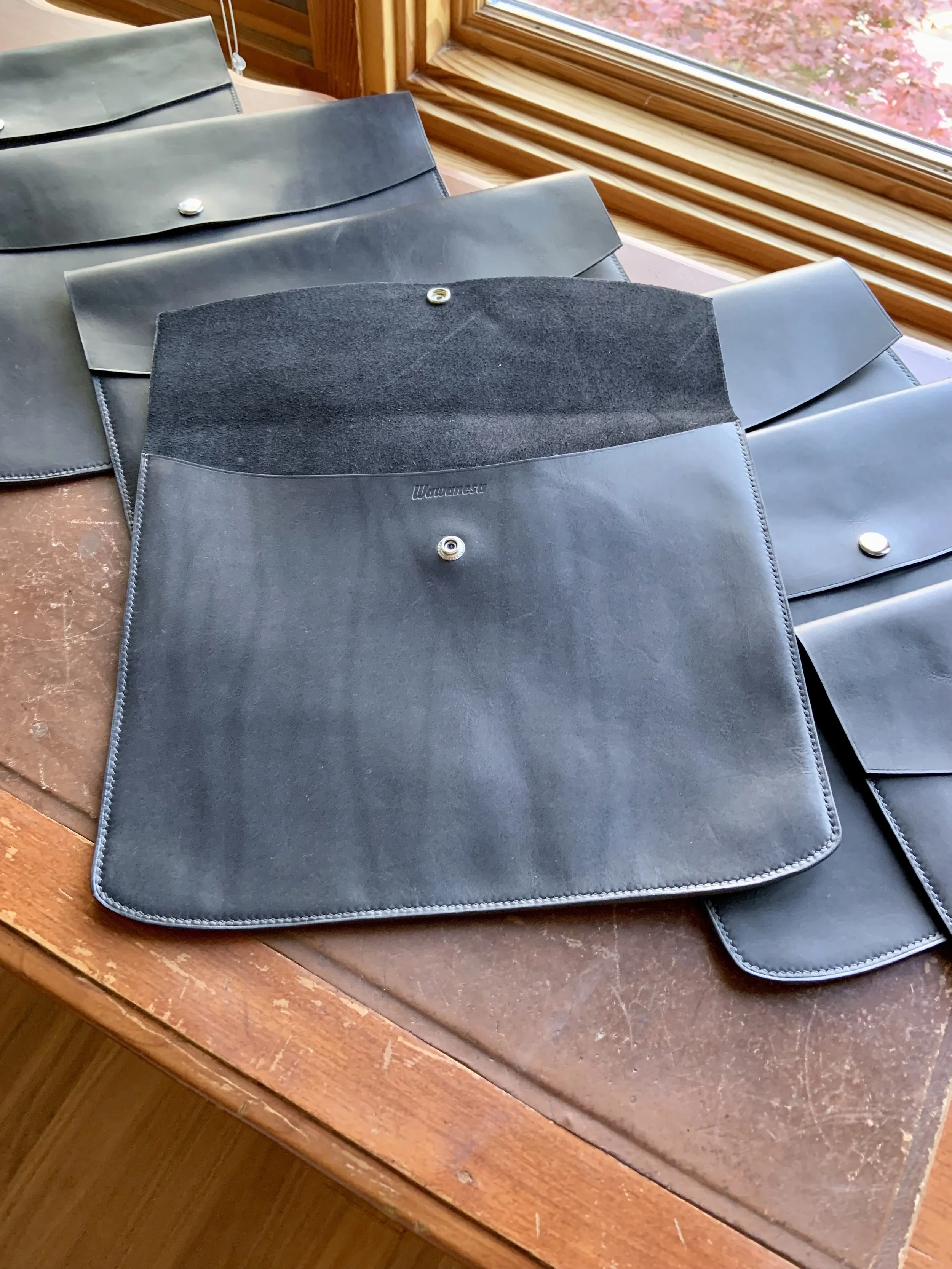 Four black leather tool pouches with snap buttons, one with a suede top, are placed on a wooden table near a window with trees outside.