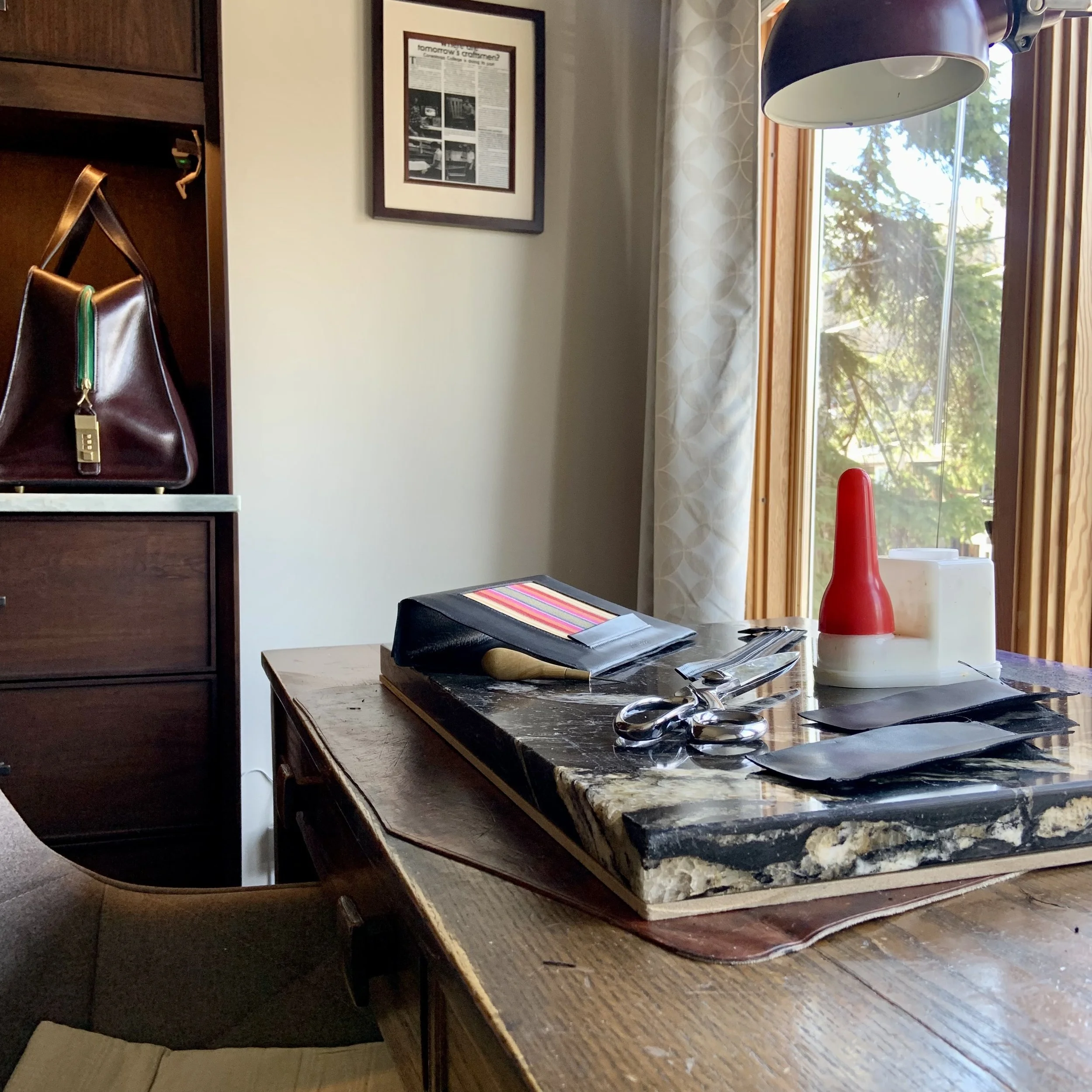 A wooden dining table with a marble top, holding a black and colorful striped folder, scissors, scissors, a red and white condiment bottle, and black napkins. To the left, a portion of a brown upholstered chair is visible. In the background, there's a window with beige curtains and an outdoor view of trees, partially obstructed by a lamp hanging from the ceiling. A framed newspaper clipping hangs on the wall.