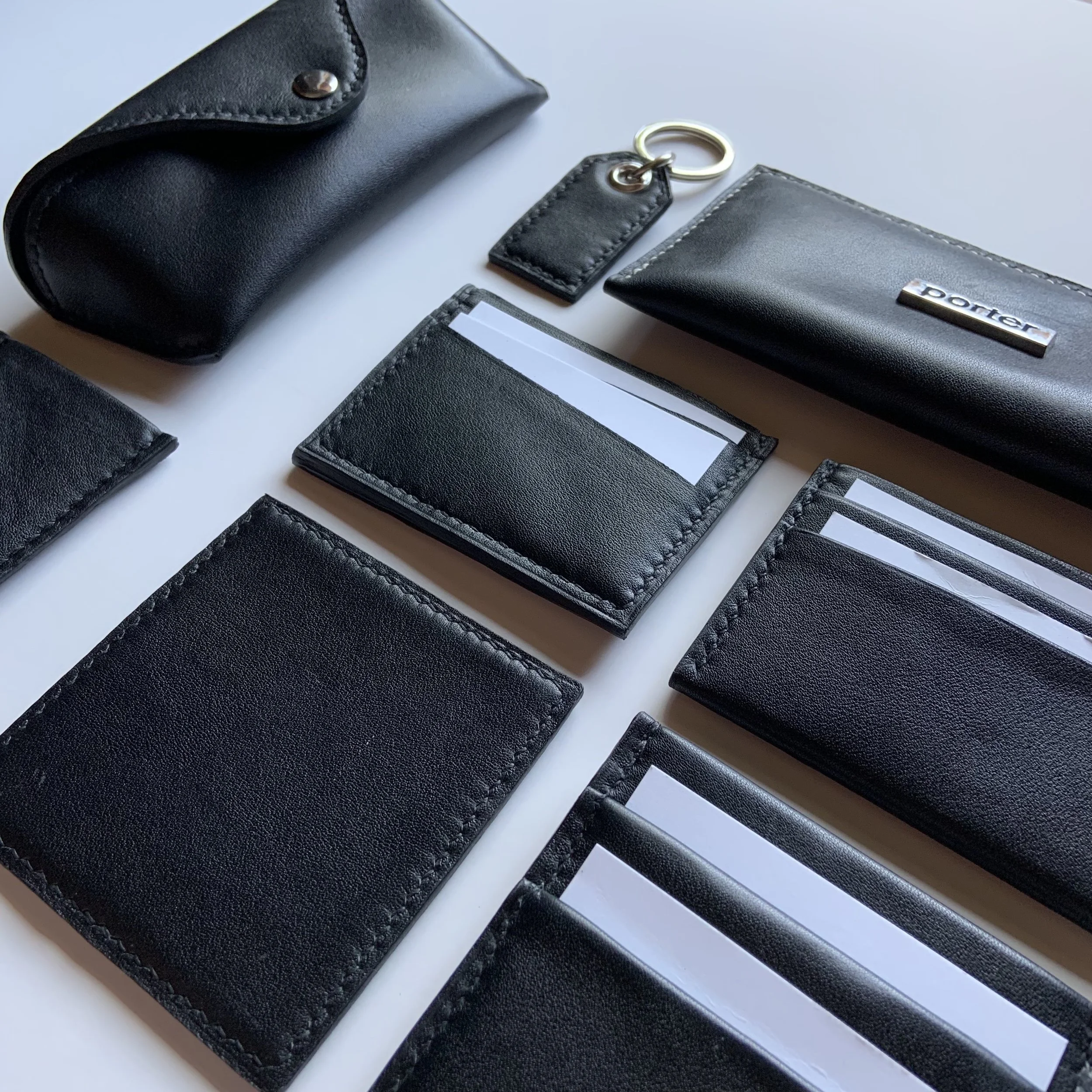 Collection of black leather accessories, including a wallet, cardholder, keyring with leather tag, and a small zippered pouch, arranged on a white surface.