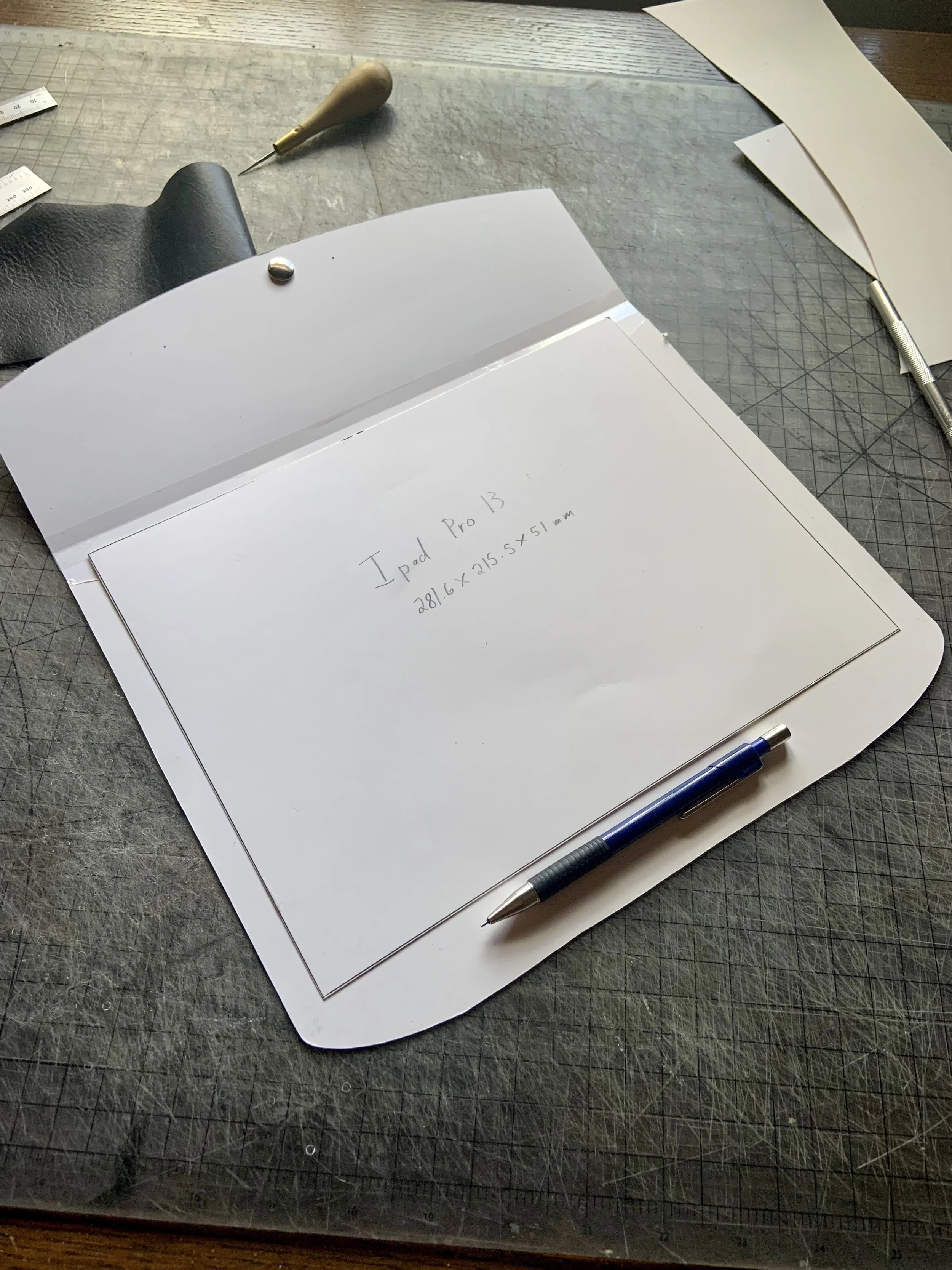 A worktable with a cutting mat, a glue stick, a curved piece of paper, a pen, and a handwritten note reading 'iPad Pro 13, 281.6 x 215.5 mm'.