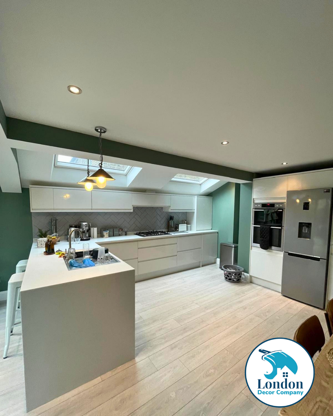 Open plan kitchen with white gloss units, grey herringbone splashback and skylights, alongside Farrow & Ball Card Room Green walls.