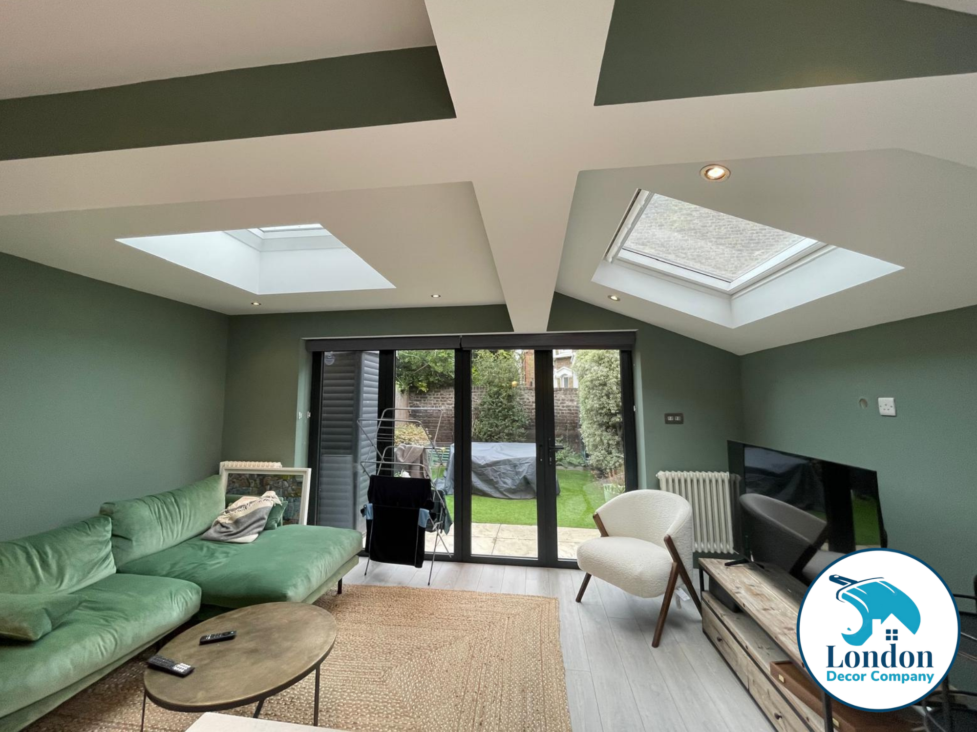 Open plan lounge with Farrow & Ball Card Room Green walls, white vaulted ceiling, skylights and green sofa facing bifold doors to the garden.