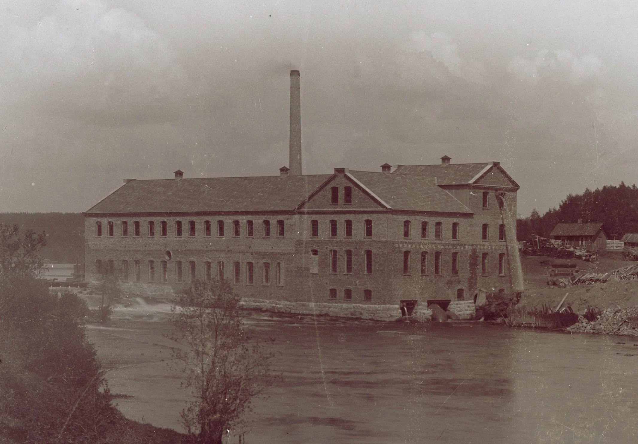 En gammal sepiafärgad foto av ett stort, industriellt tegelhus med många fönster, en hög skorsten i bakgrunden och en flod framför byggnaden.