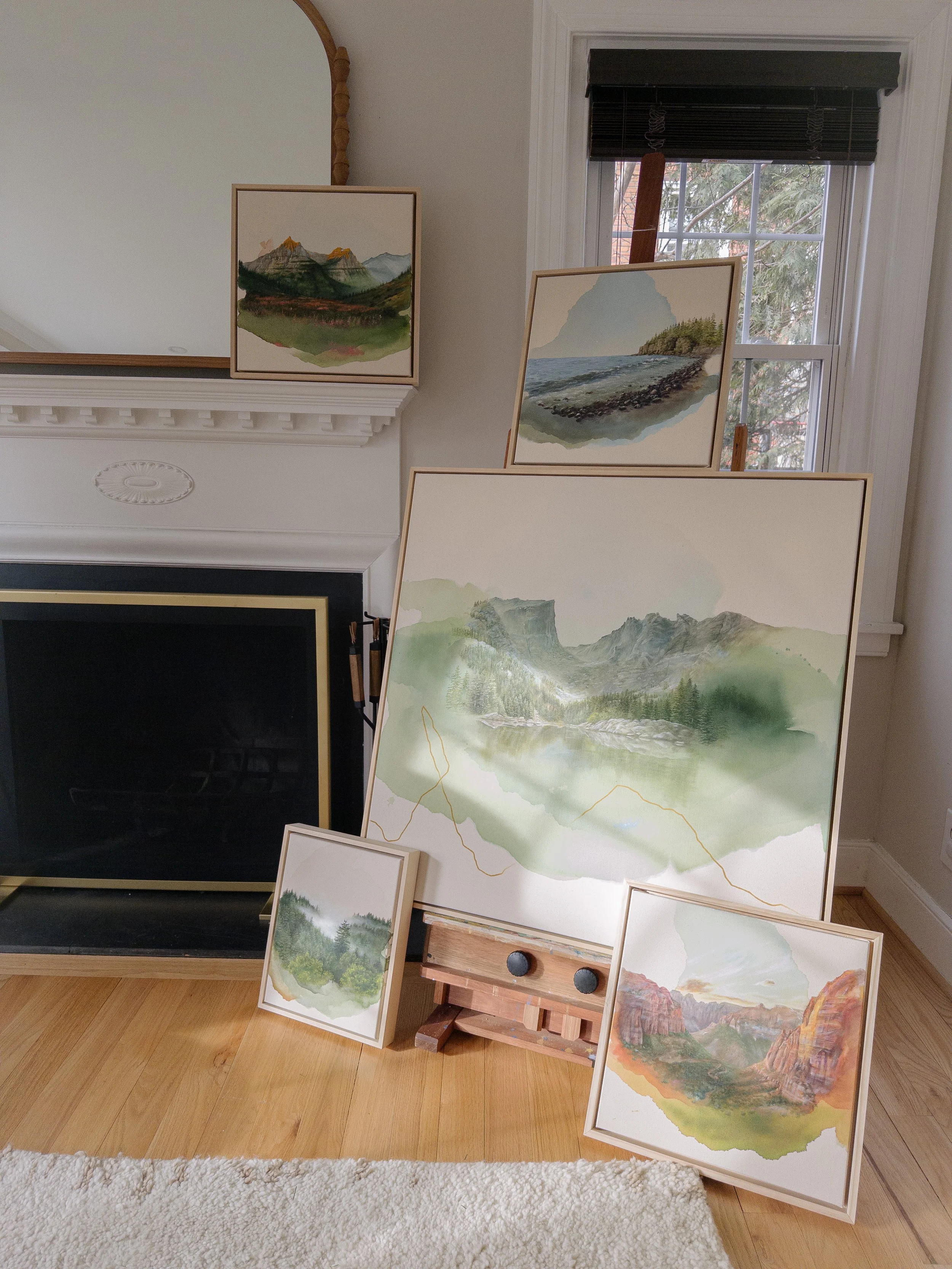 A collection of five landscape watercolor paintings on display in a room, with some leaning against a fireplace and others on an easel, featuring mountains, lakes, and trees.