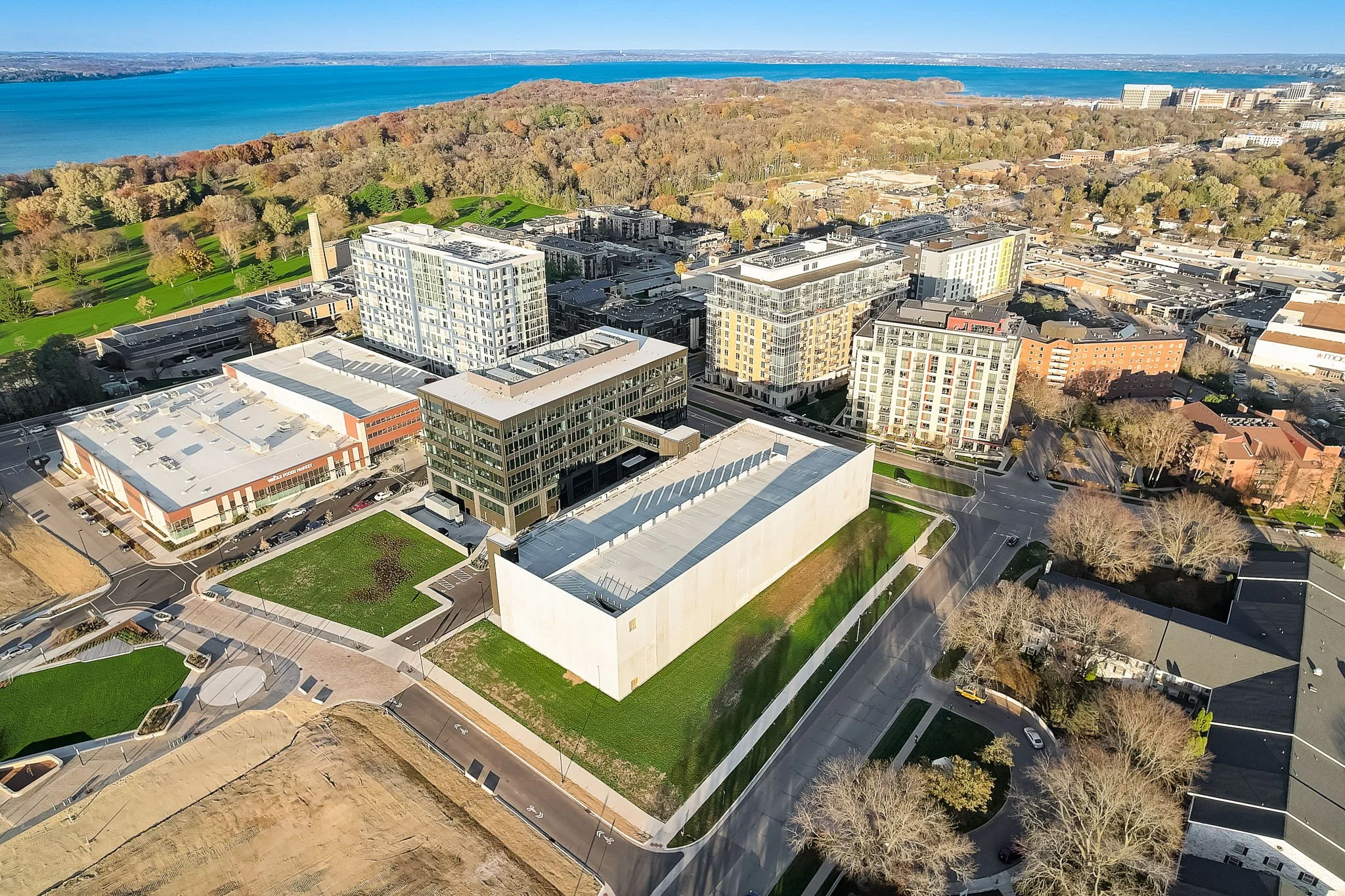 Aerial view of a cityscape with modern buildings, green spaces, and a large body of water in the background.