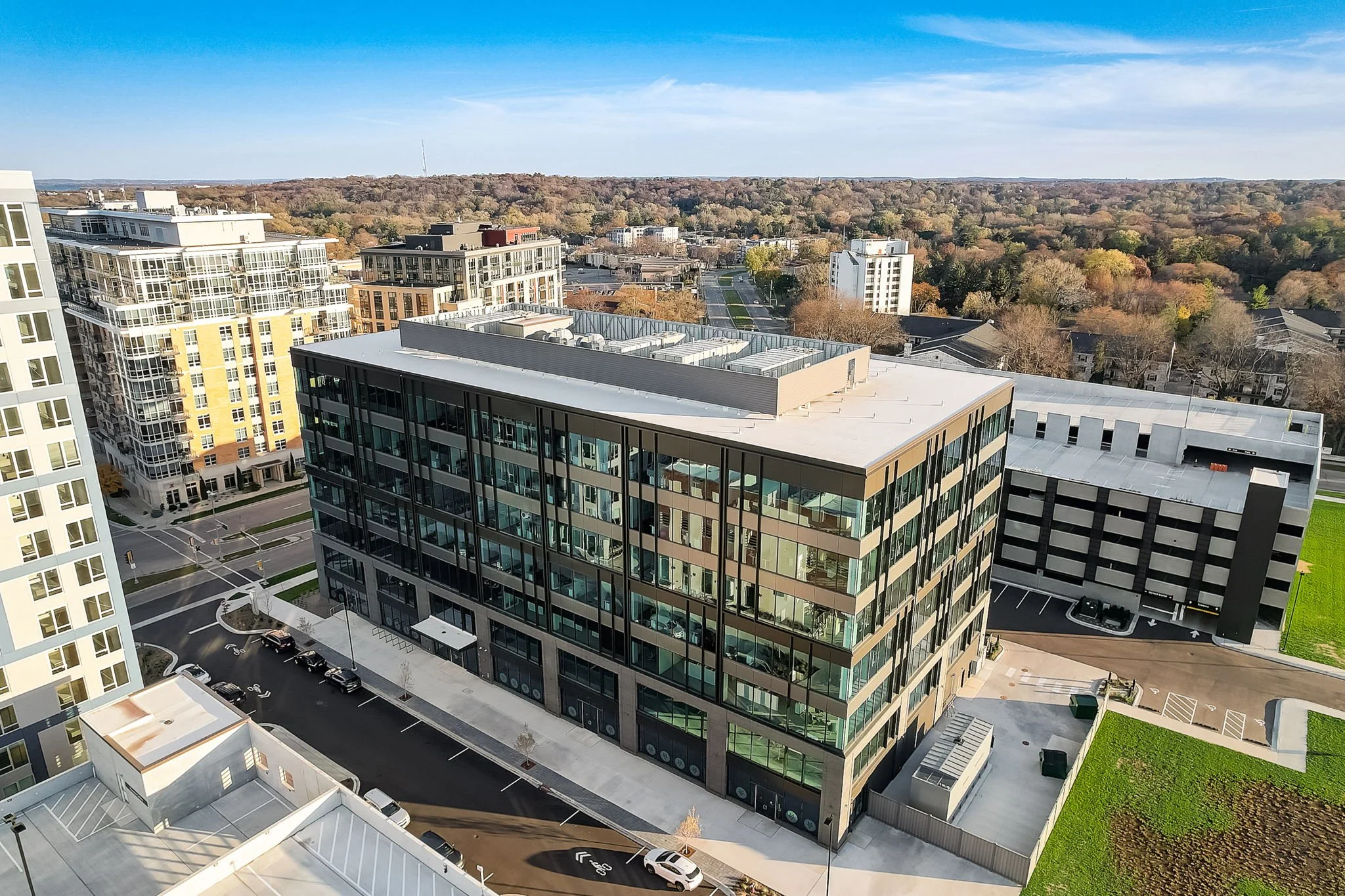 Aerial view of a modern urban area with multiple multi-story buildings, including a glass and metal office building, other residential and commercial structures, and some greenery. The scene is under a clear blue sky.