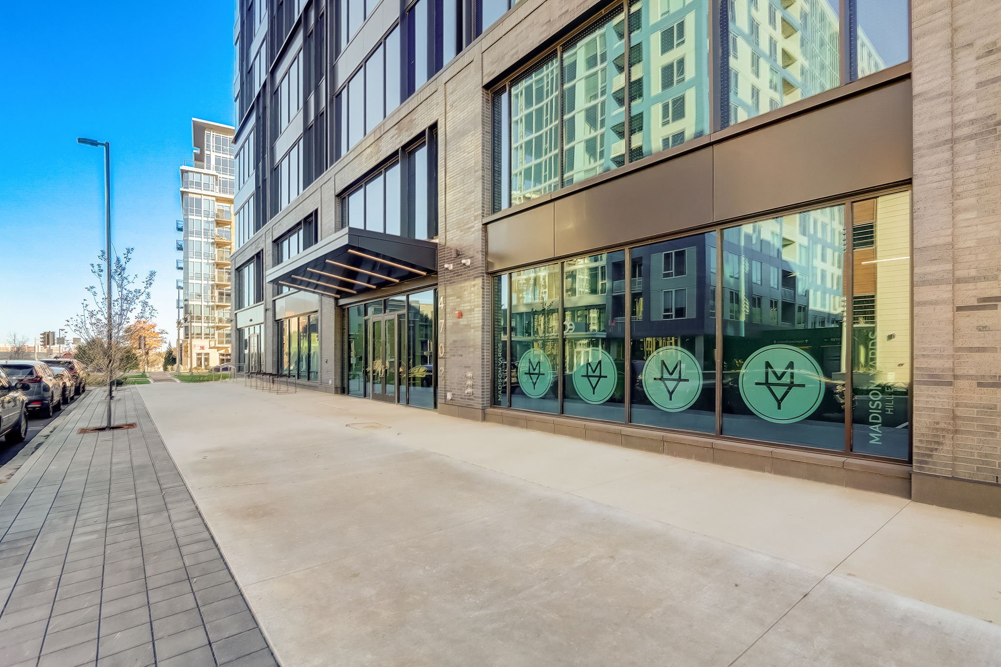 Modern urban building with glass facade and sidewalk, cars parked alongside, clear sky in the background.