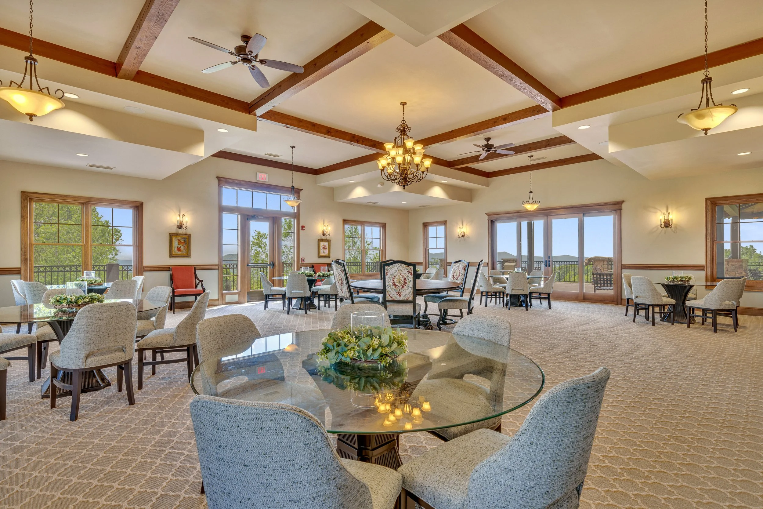 Interior of a spacious dining or event room with large windows and outdoor views, decorated with chandeliers, ceiling fans, and a mix of round and rectangular tables with chairs, some with floral arrangements.