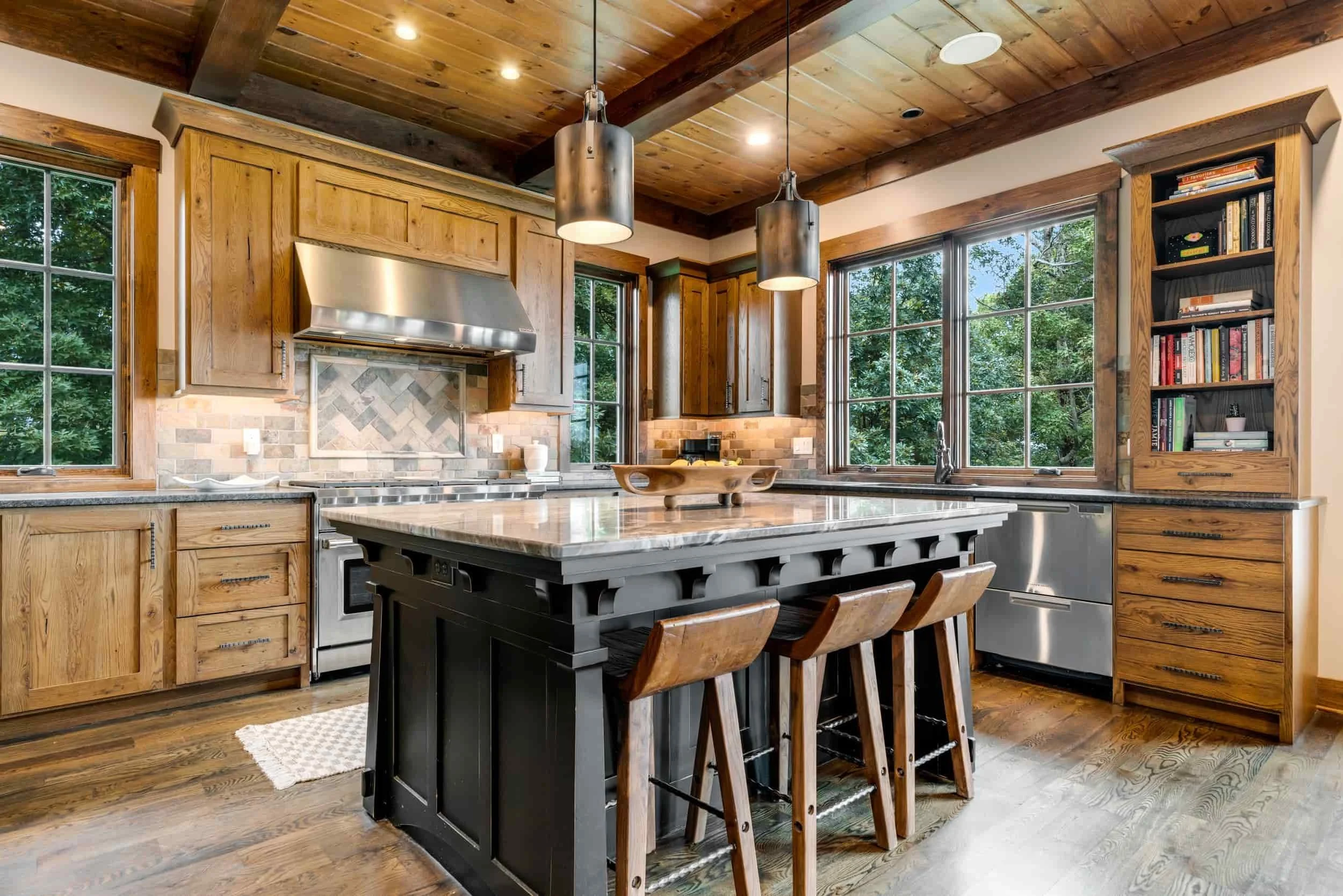 A luxury mountain home kitchen in Grand Highlands at Bearwallow Mountain Hendersonville NC with wooden cabinetry, a large central island with a marble countertop, and three modern pendant lights.