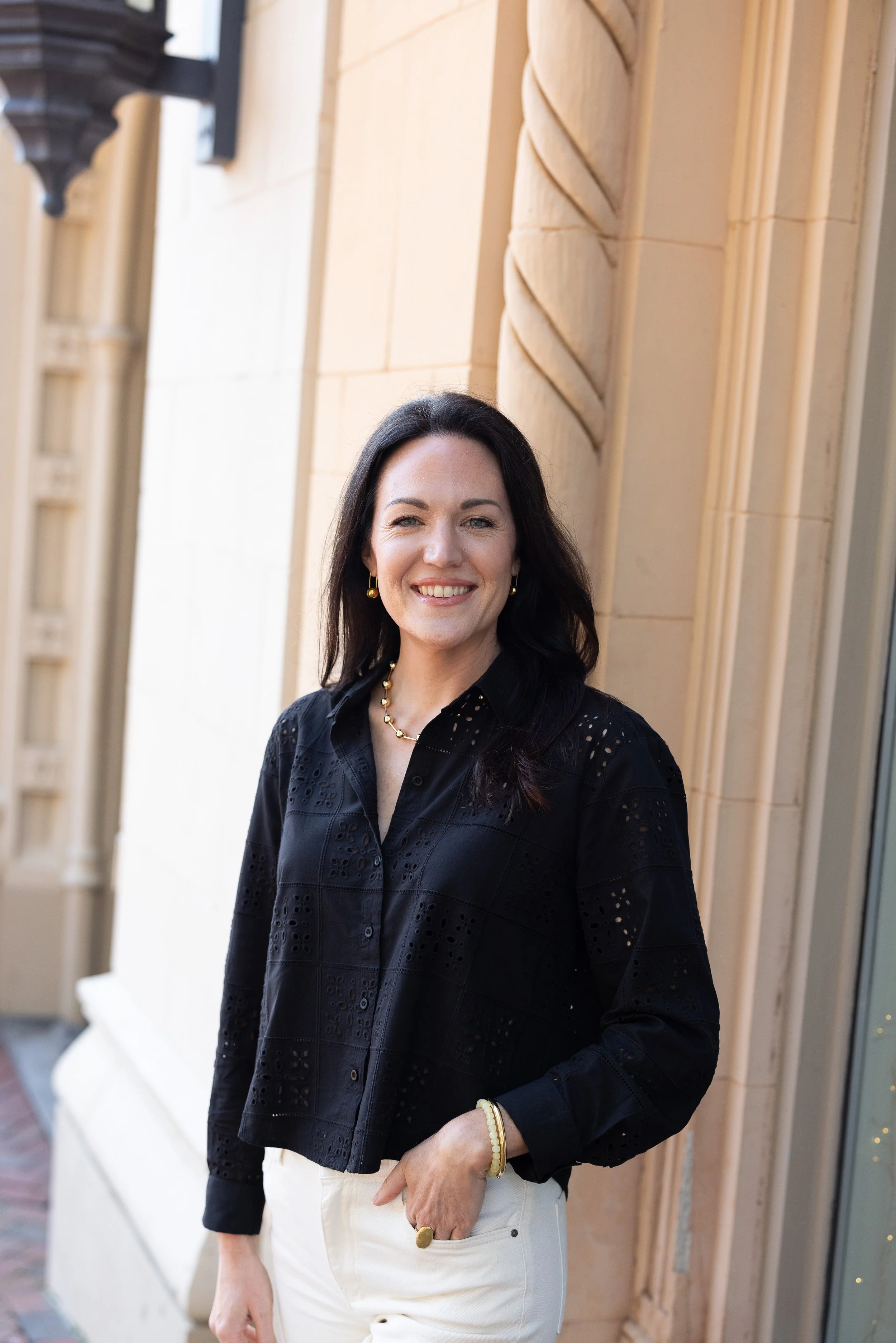 Ashton Peters Realtor smiling outdoors in front of a beige stone building, wearing a black embroidered blouse, white pants, and gold jewelry.